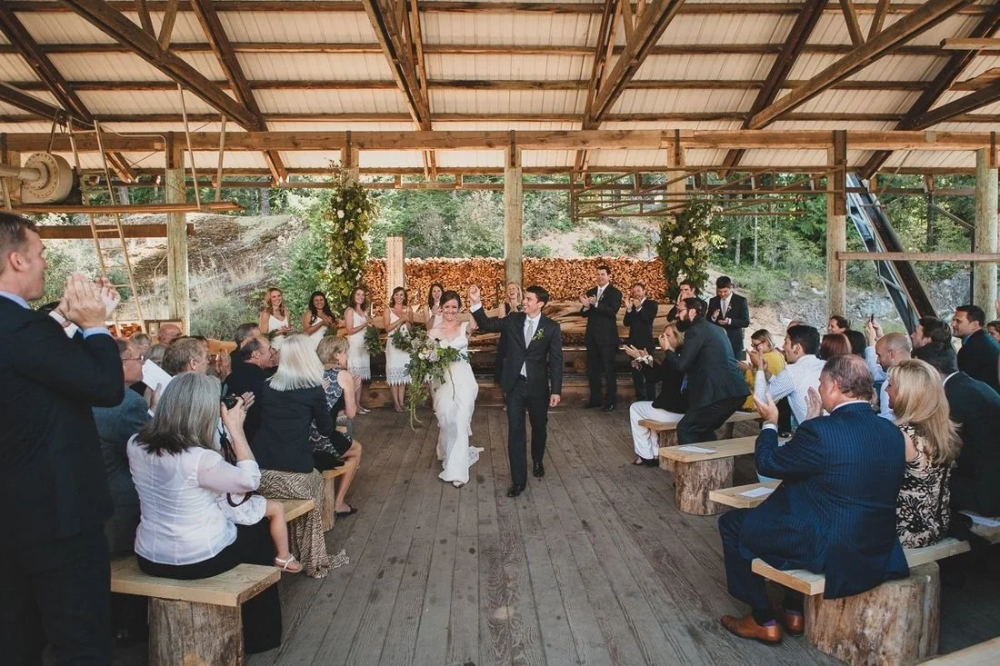 Bride and groom walking and waving in a rustic wedding ceremony with seated guests and a wooden backdrop with flowers and logs.