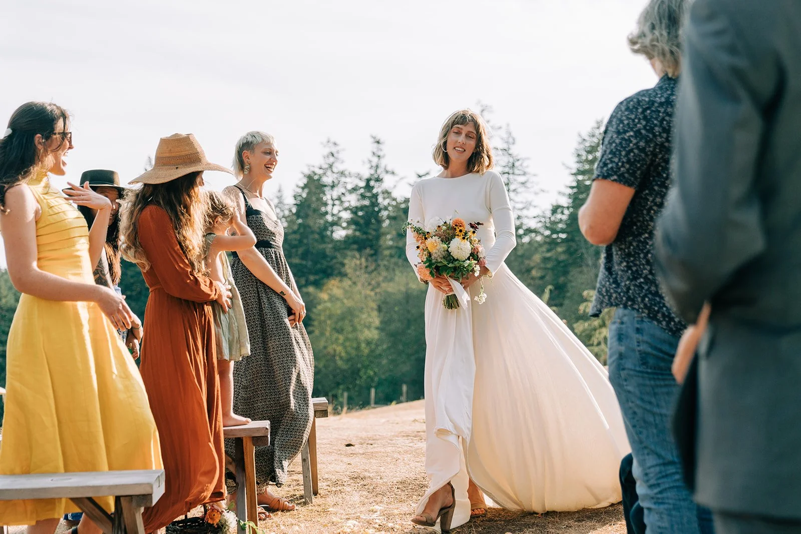 A woman in a white dress holding a bouquet of flowers at an outdoor wedding ceremony with guests around her.