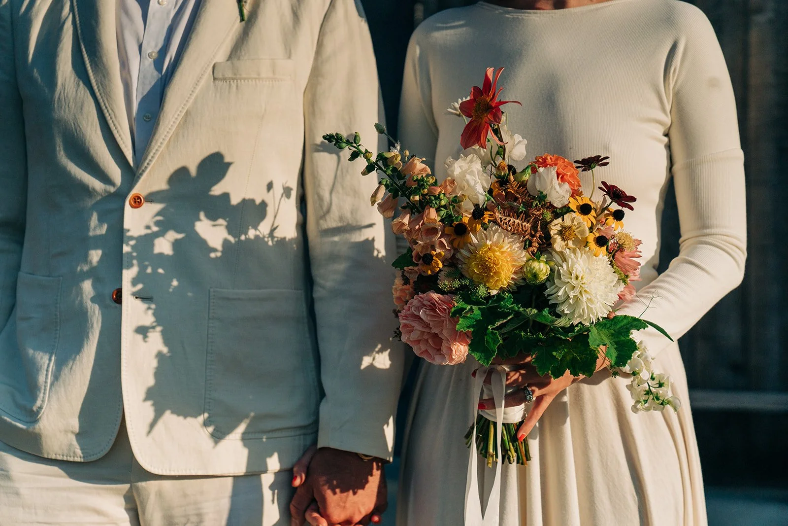 Close-up of a couple holding hands, with the woman holding a bouquet of various flowers, both dressed in wedding attire.