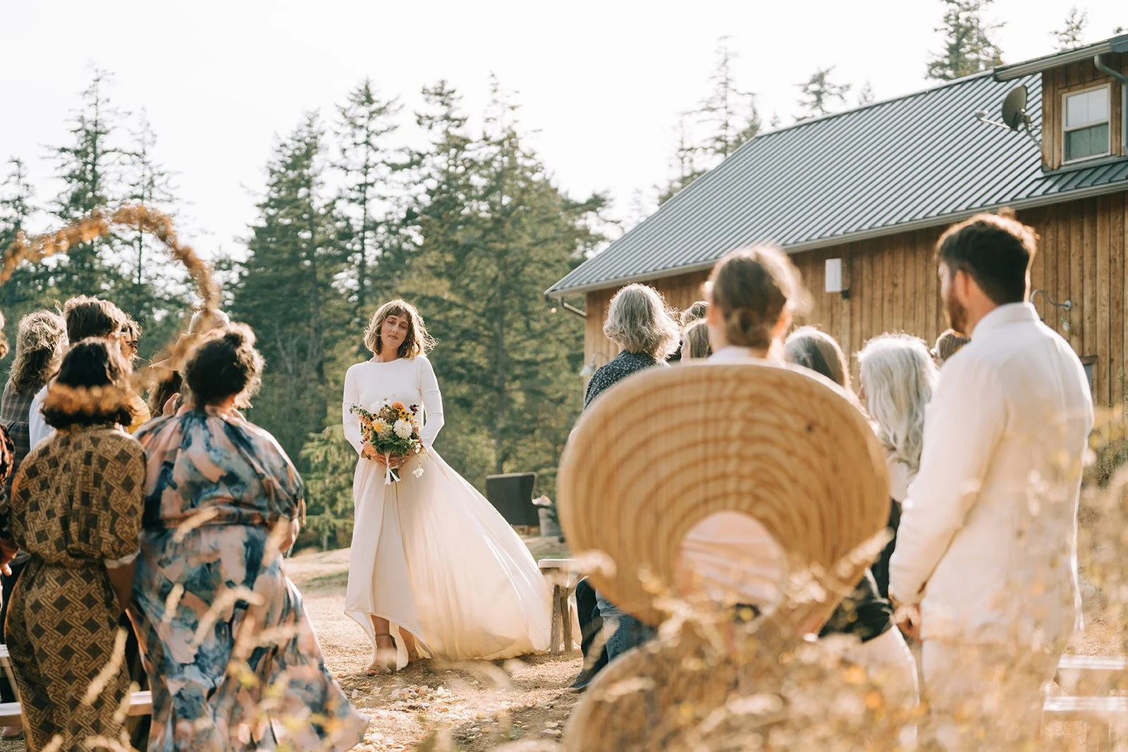 A bride in a white wedding gown holding a bouquet of flowers, standing outdoors during a wedding ceremony with guests seated on wooden benches, surrounded by trees and a wooden building in the background.