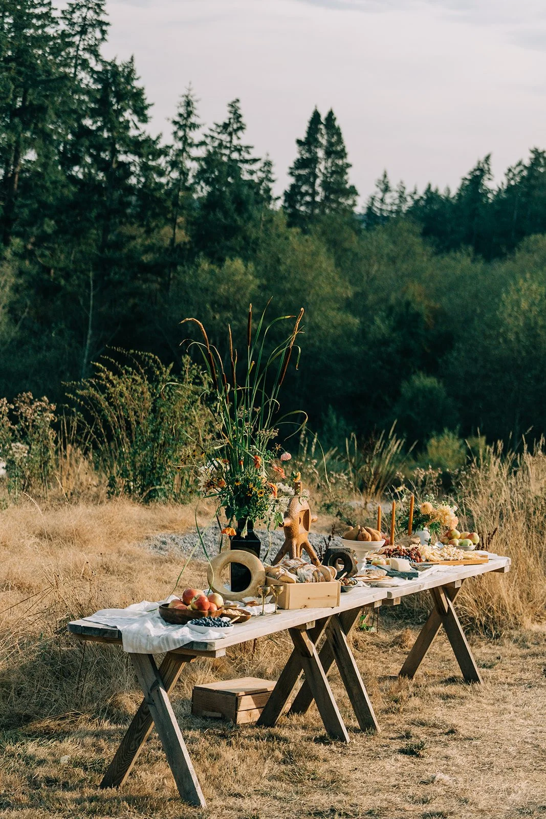 A rustic outdoor dining table set in a grassy field with a forest backdrop, decorated with flowers, candles, and various food items such as fruits, bread, and cheese.