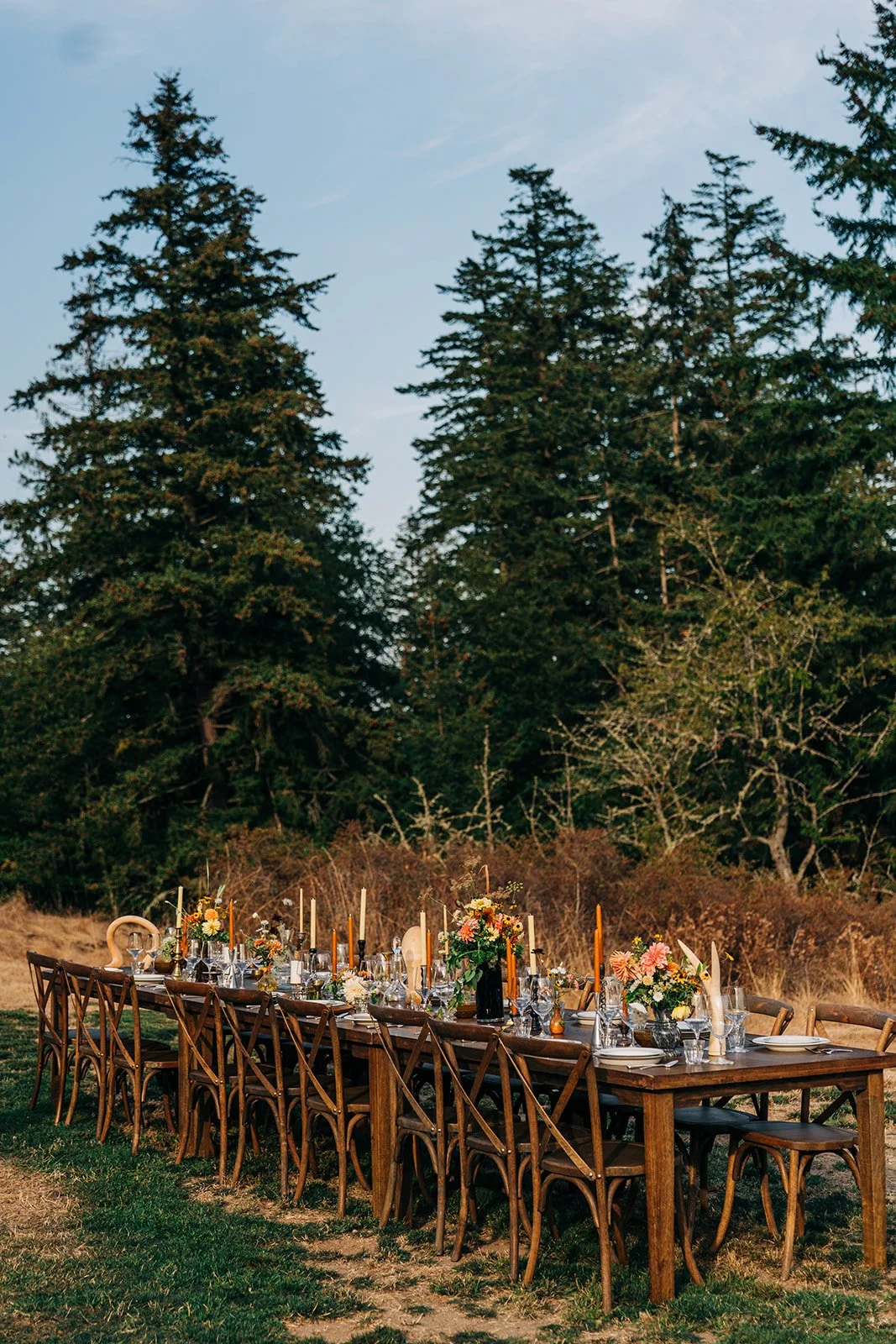 Outdoor dining table set with floral centerpieces and candles in a natural forest setting.