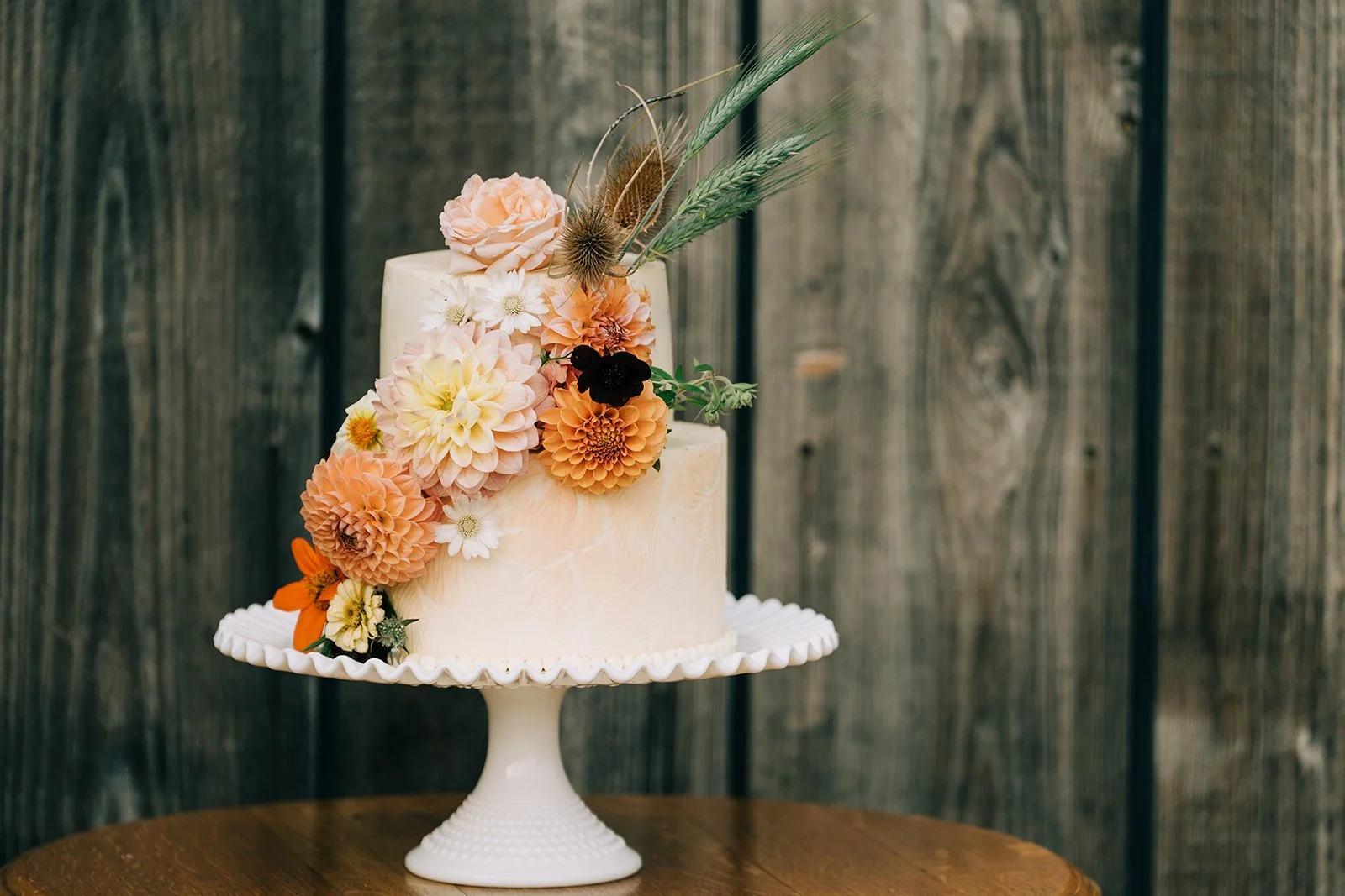 A two-tiered wedding cake decorated with fresh flowers and greenery, placed on a white cake stand on a wooden table against a rustic wooden fence background.