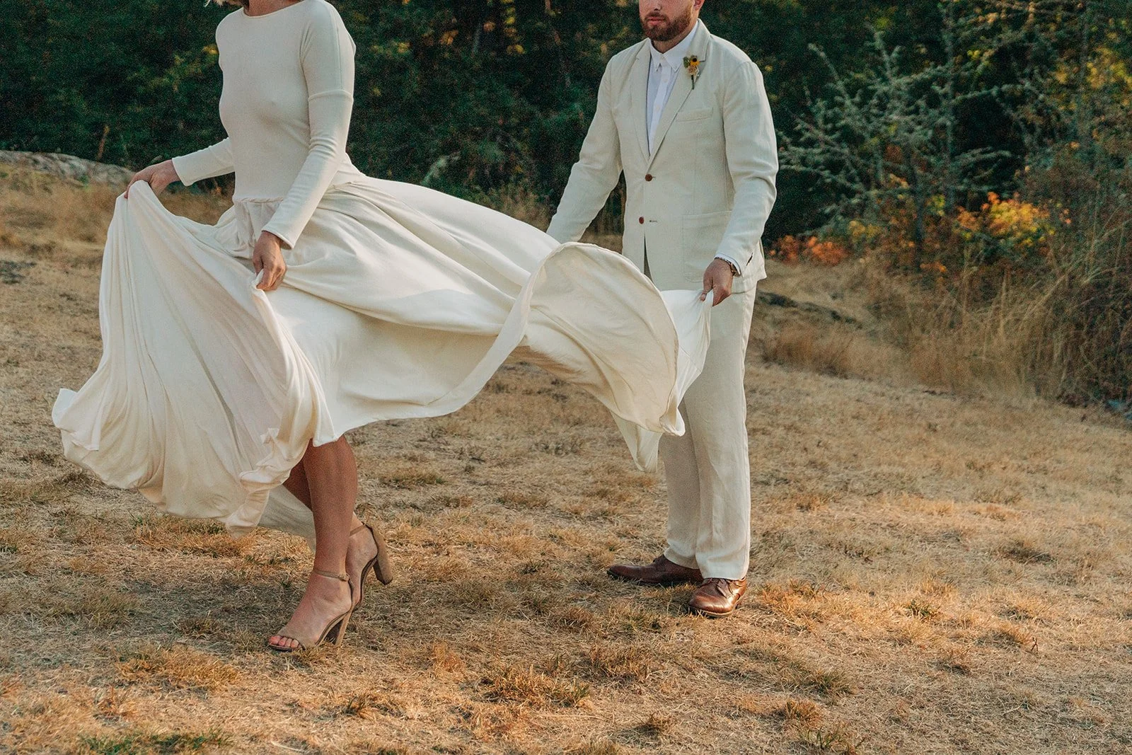 A couple in wedding attire standing outdoors on dry grass in a natural setting. The woman is wearing a long, flowing white dress and high heels, and the man is in a cream suit. They are holding and lifting the woman's dress, revealing her legs and sh