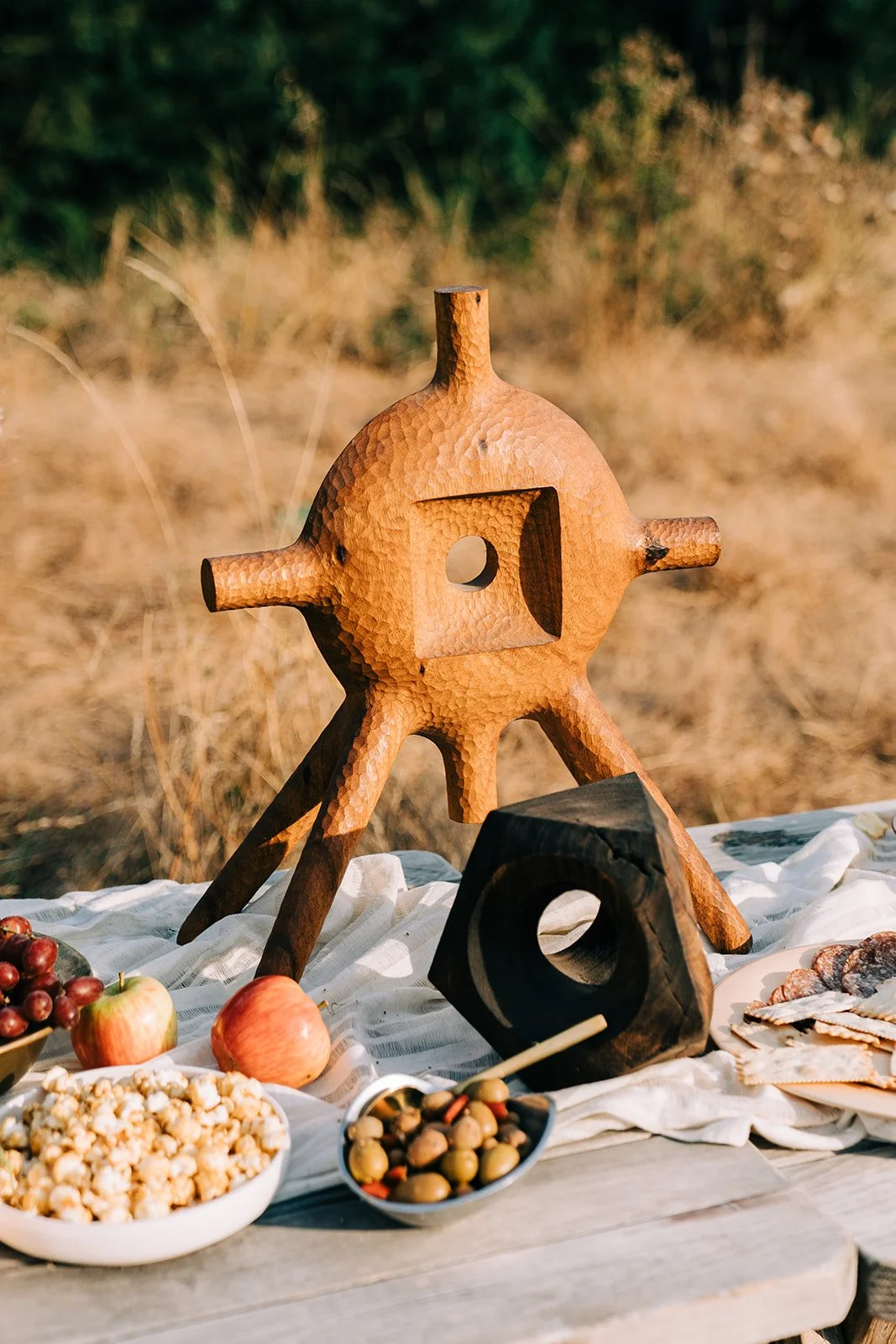 A rustic outdoor table set with snacks and a wooden cheese board sculpture in the shape of a gear, with a background of dry grass and greenery.