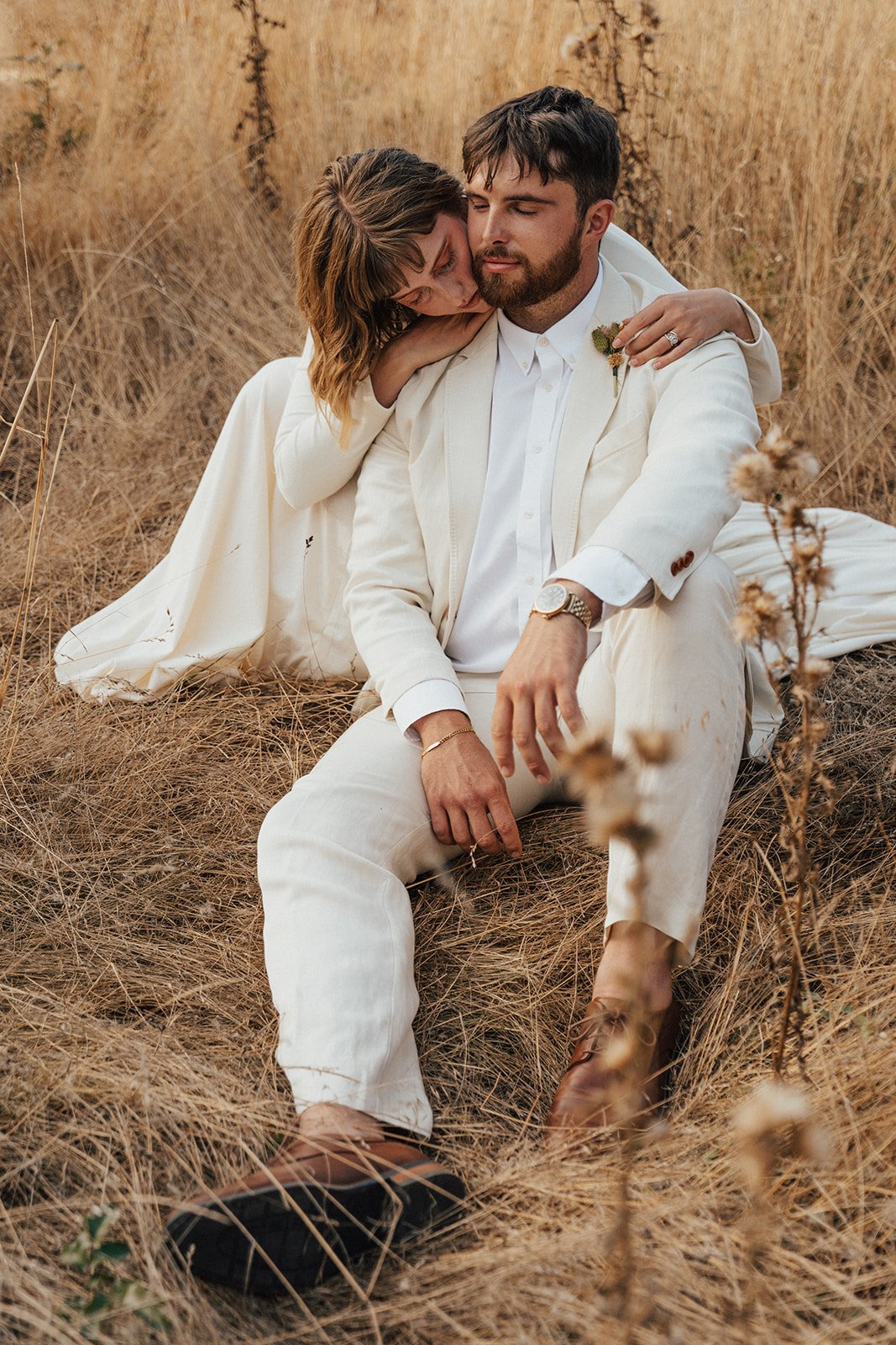 A couple dressed in white sitting in a dry grassy field, with the woman leaning on the man's shoulder.