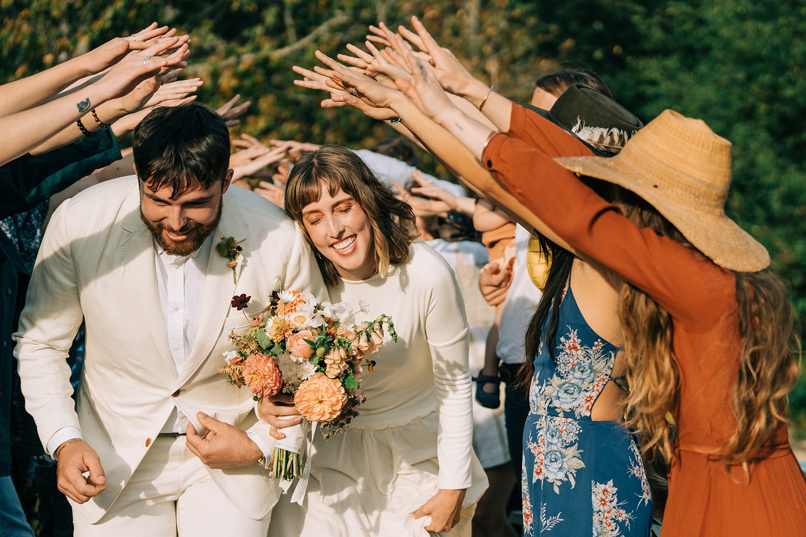 A joyful bride and groom leaning, smiling, under a canopy of raised hands, during a celebration outdoors with green trees in the background.
