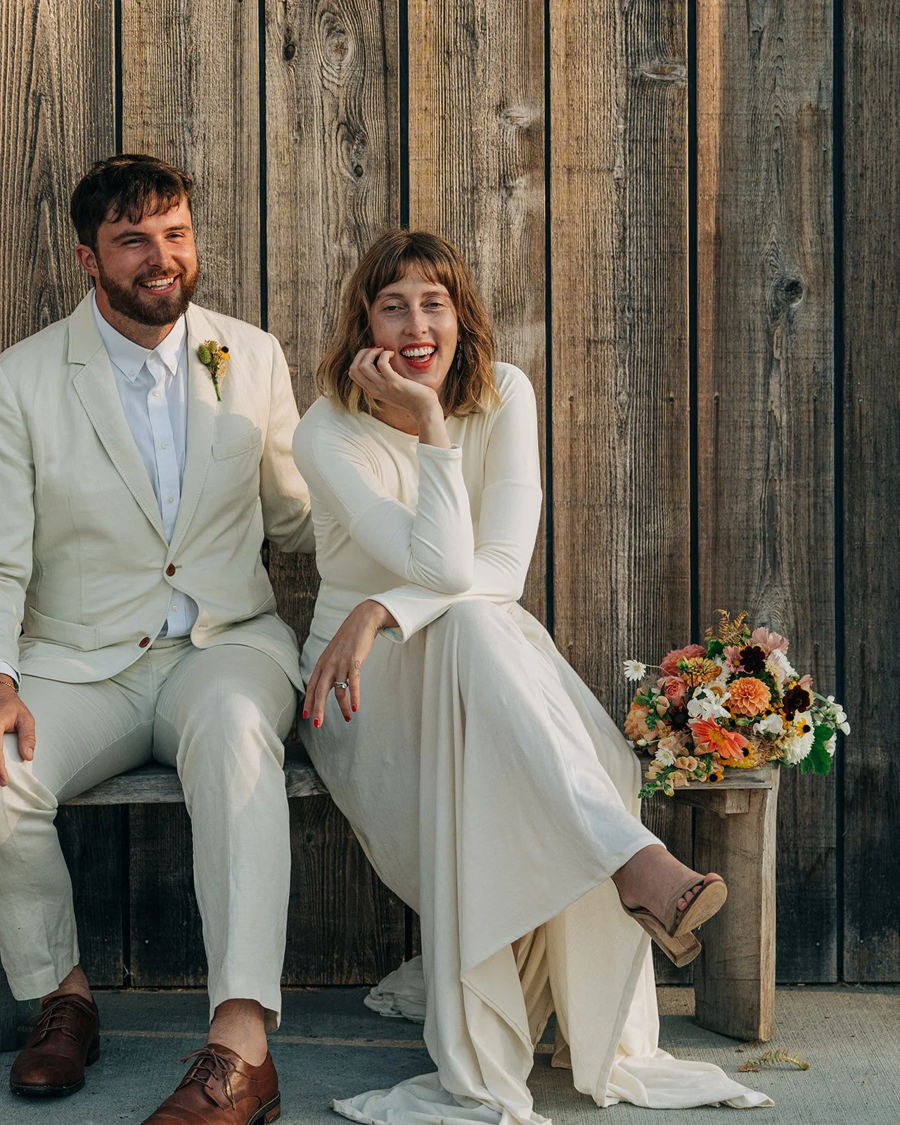 A man and woman dressed in white sitting on a wooden bench beside a bouquet of flowers, with a wooden wall in the background.