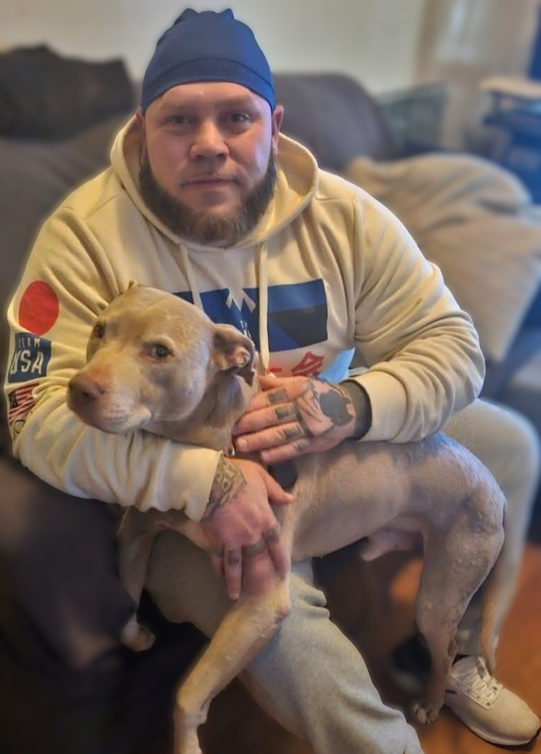 A man with a beard, tattoos, and wearing a blue head covering and beige hoodie with patches, sitting on a couch and holding a large tan dog with a muscular build and light eyes in his lap.