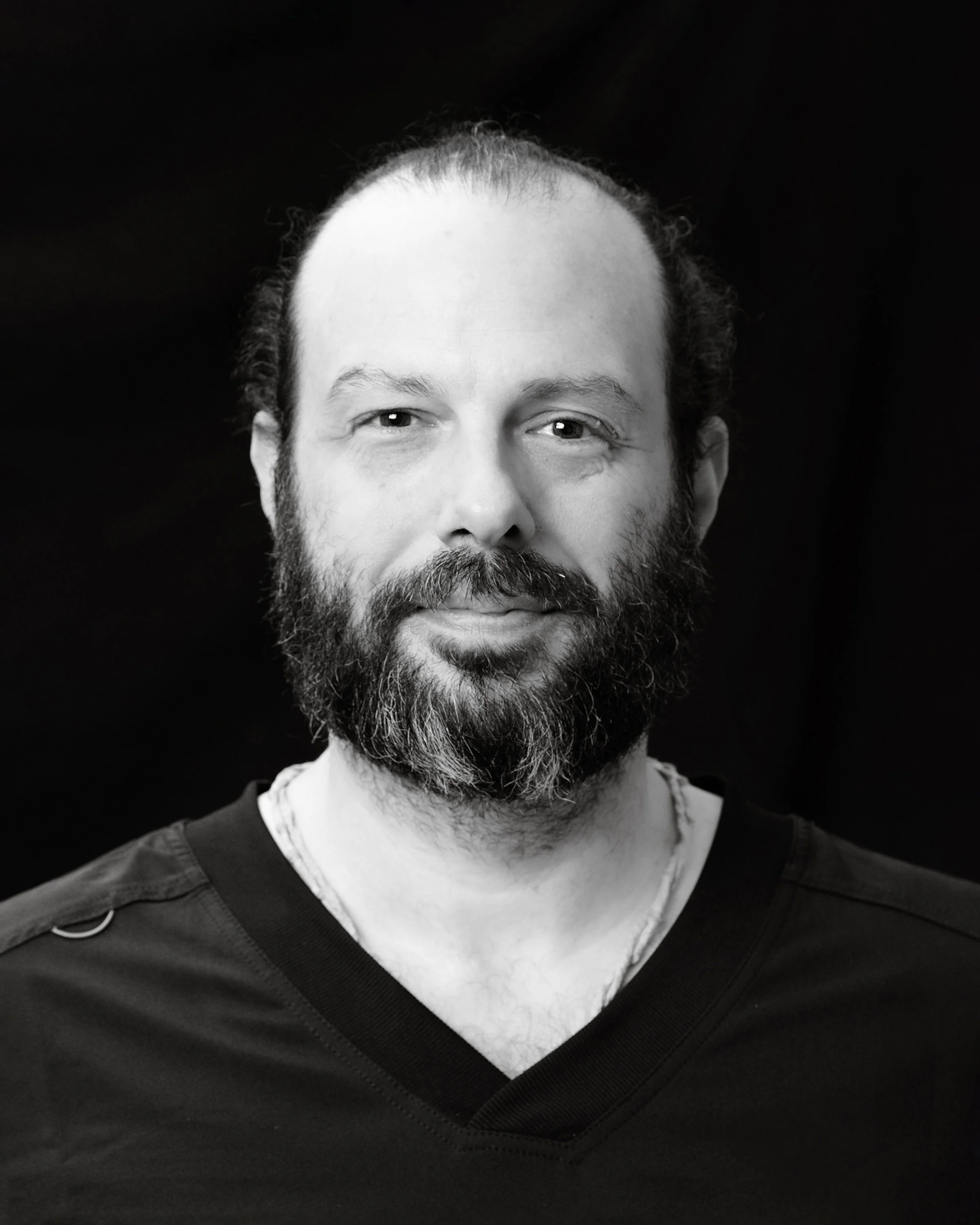 Black and white portrait of a woman with wavy hair, light eyes, and a small nose piercing, wearing a black V-neck shirt, against a dark background.