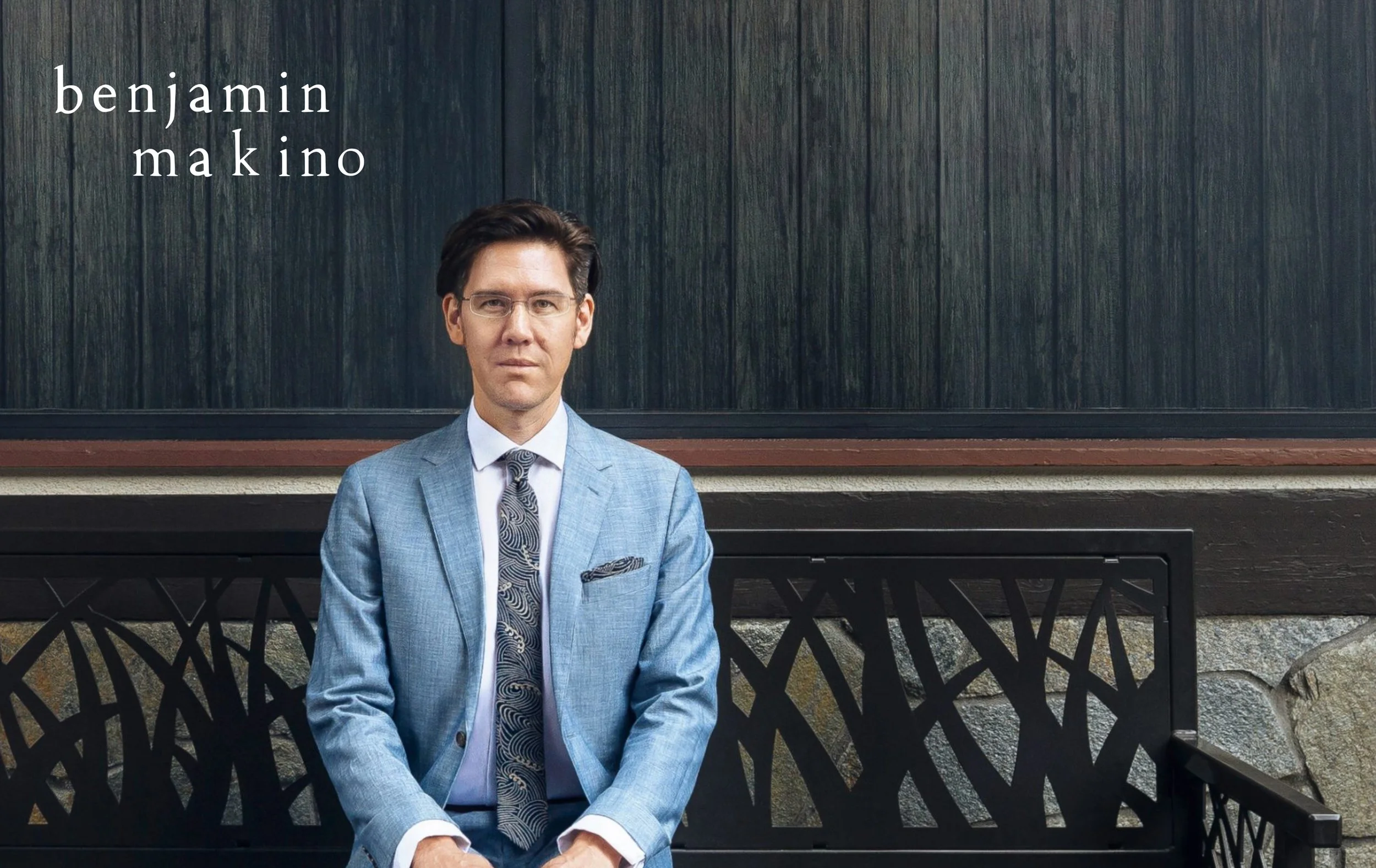 A man wearing glasses in a blue suit and patterned tie sitting on a decorative black metal bench with a stone base, in front of a dark green wooden wall with text overlay that reads 'benjamin makino'.