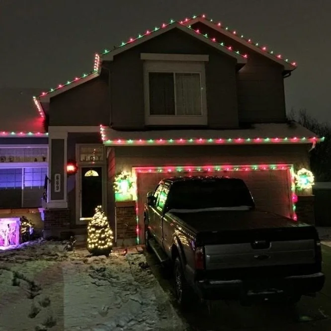 A two-story house decorated with multicolored Christmas lights on the roofline, garage, and porch. A black pickup truck is parked in the driveway, and snow covers the ground. Small lit Christmas trees are placed outside near the house.