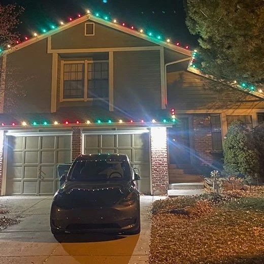 A two-story house decorated with colorful Christmas lights along the roofline. A black car is parked in the driveway in front of two garage doors. A tree and some shrubs are visible on the right side.