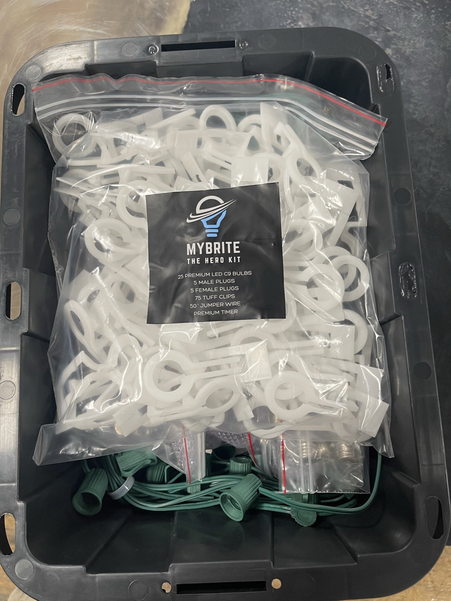 A black plastic bin containing a large sealed plastic bag of white Christmas light clips labeled 'MYBRITE THE HERO KIT,' which includes LED bulbs, plugs, clips, jumper wire, and a timer.