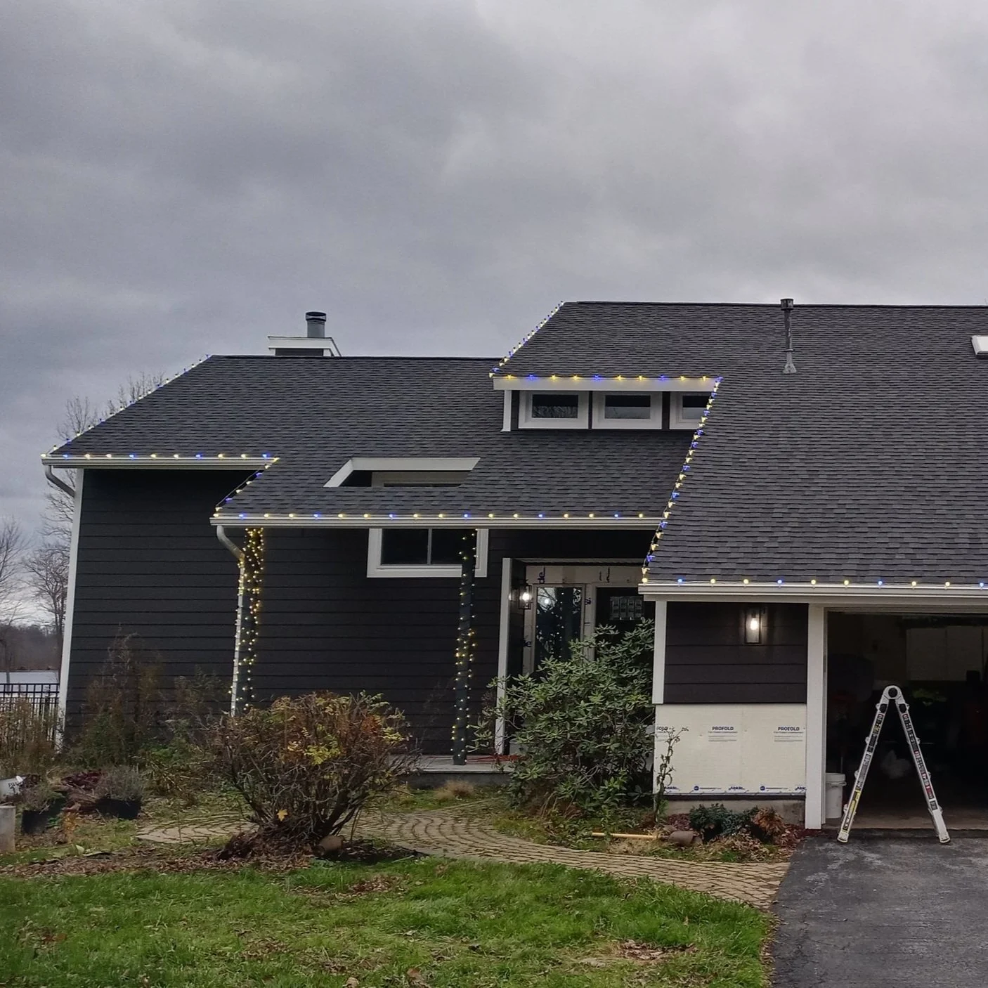 House with dark siding decorated with string lights on the roof and columns, overcast sky, front yard with bushes, driveway, and a ladder near the garage.