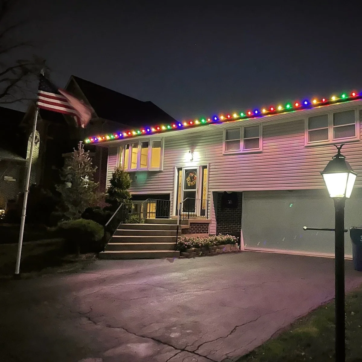 A two-story house decorated with colorful Christmas lights along the roofline, with an American flag on a flagpole in the front yard, a well-lit front porch with a wreath on the door, and landscaping including small trees and flowers. A street lamp illuminates the driveway at night.