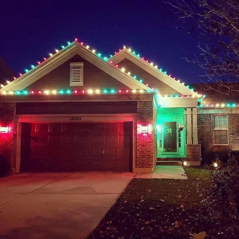 A house decorated with colorful Christmas lights, including red, green, yellow, and blue, outlining the roof and around the front porch at night.