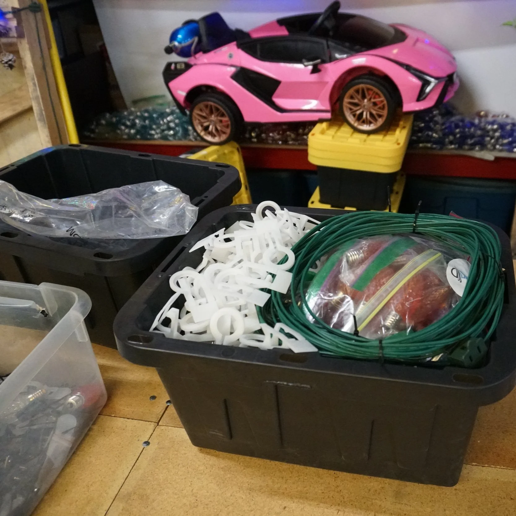 A pink toy Lamborghini sports car model with a blue helmet on top, placed on yellow storage bins, surrounded by plastic containers filled with various small parts, tools, and wires in a workshop or garage setting.