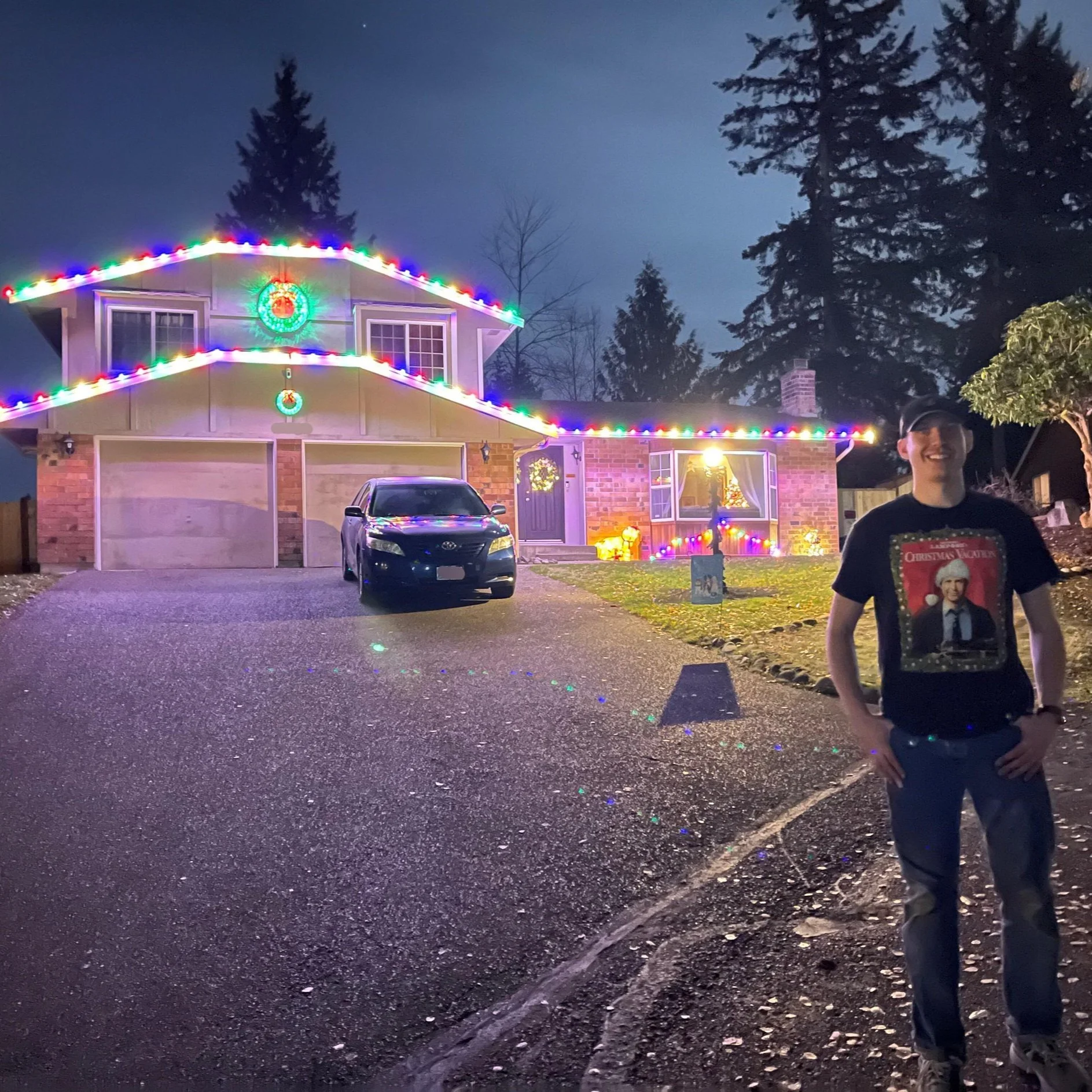A house decorated with colorful Christmas lights at night, with a man standing on the driveway smiling, wearing a black t-shirt with a Christmas-themed graphic. The house has illuminated wreaths and decorations, and a black car is parked in front.