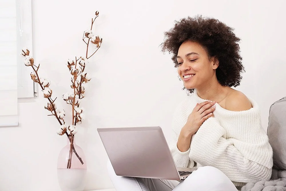 A woman with curly hair smiling while sitting on a couch with a laptop, in a bright room with a cotton branch in a vase on the wall.