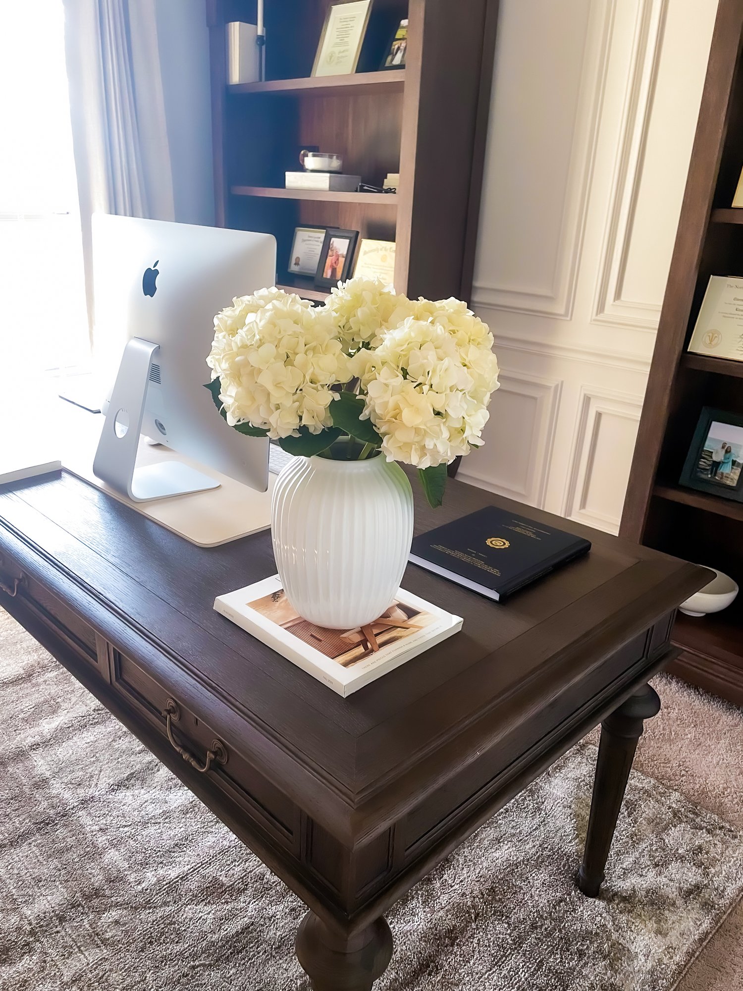 A dark wooden desk holding a white vase with white hydrangea flowers, a closed black notebook, and a magazine, with an iMac computer in the background, in a room with bookshelves and framed pictures.
