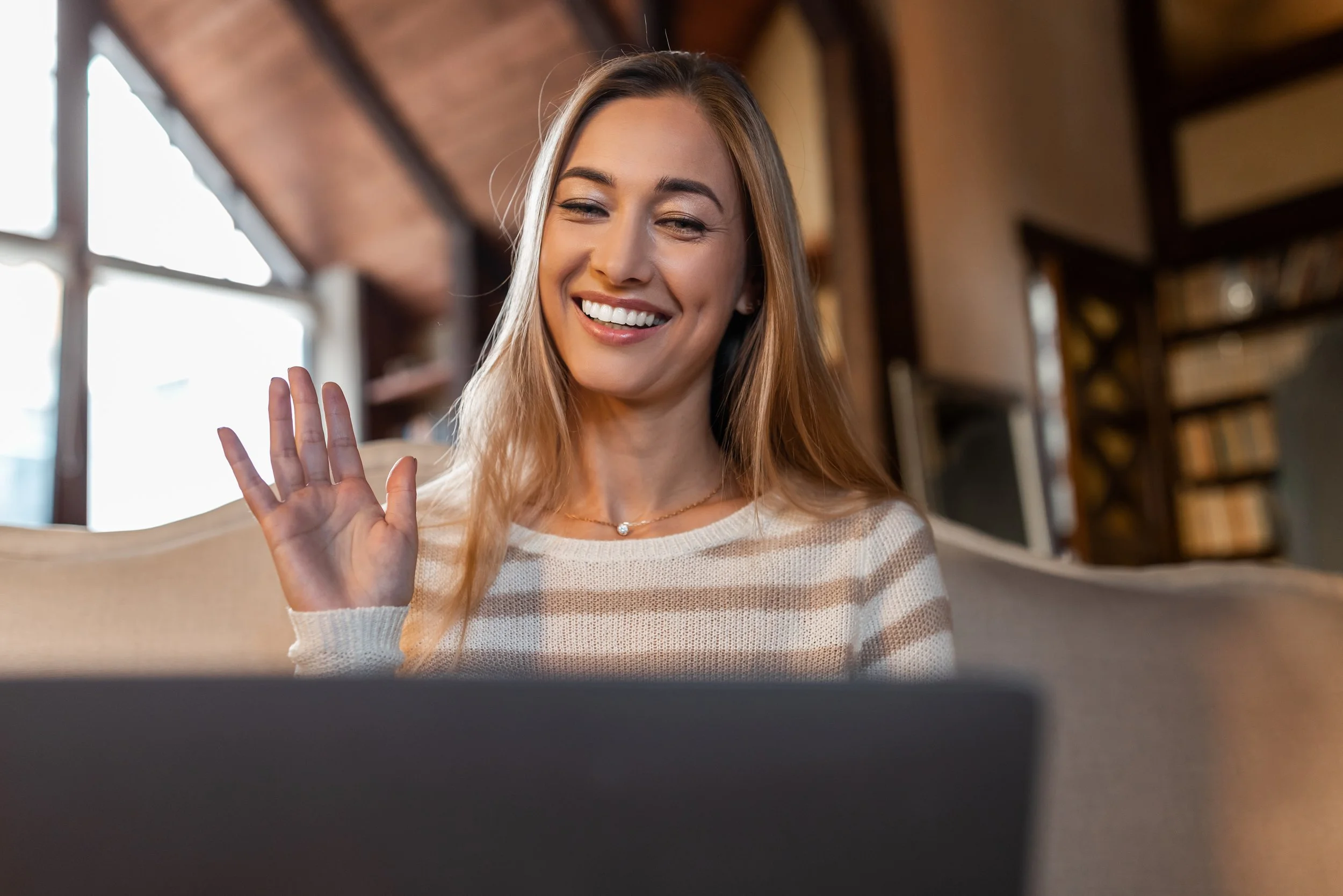 Young woman with long blonde hair smiling and waving at her laptop in a cozy room with wooden ceiling and shelves in the background.