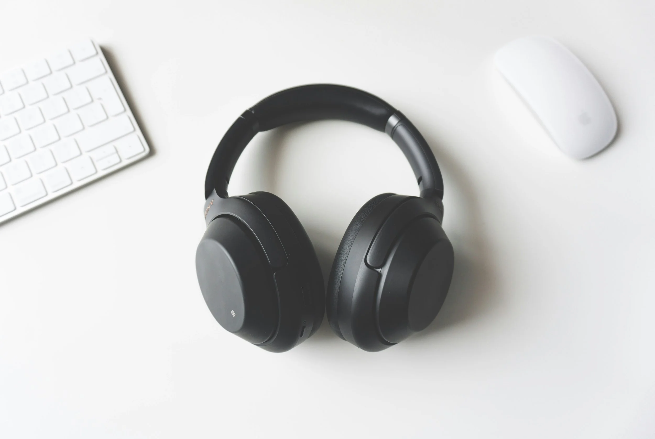 A pictture of black headphones on a white desk, with a white keyboard and a white mouse.