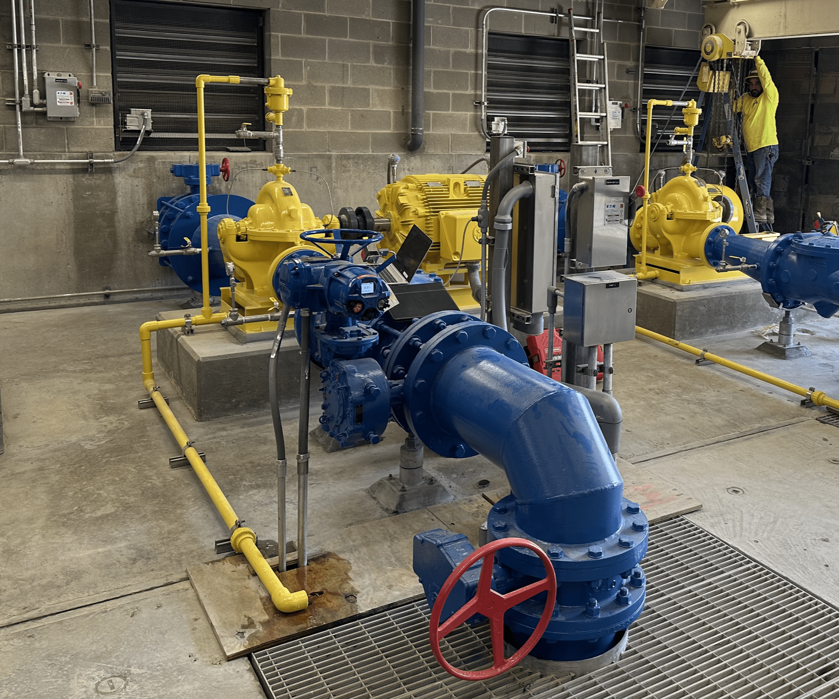 Industrial pump system with blue and yellow components in a utility room, man in yellow jacket standing on a ladder