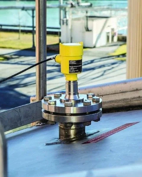 A yellow industrial sensor mounted on a metal flange connected to a pipe or machinery in an outdoor industrial setting.