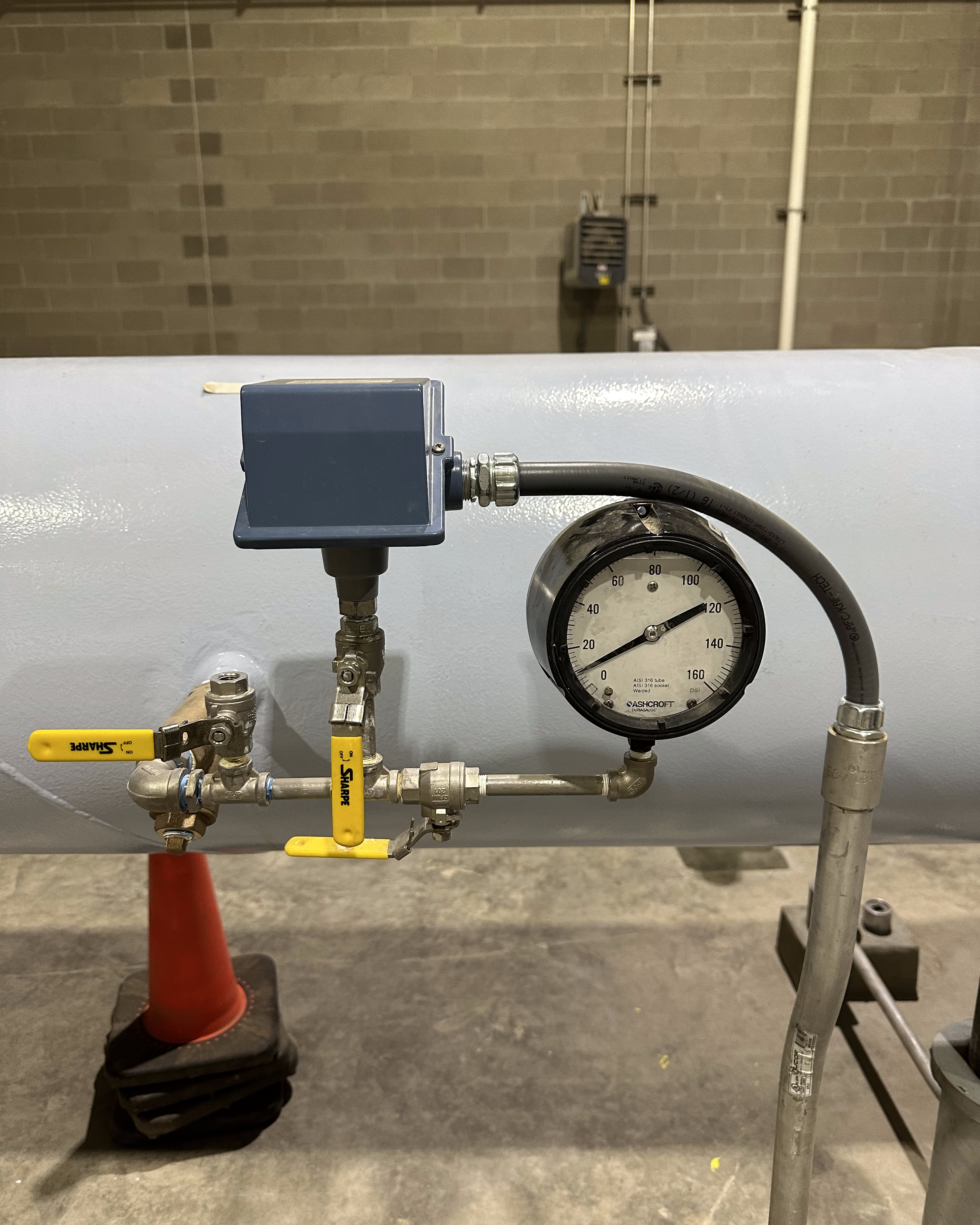 Close-up of a pressure gauge and piping on a white industrial machine with yellow valves, in a room with brick walls and concrete floor, partially obscured by a red safety cone.