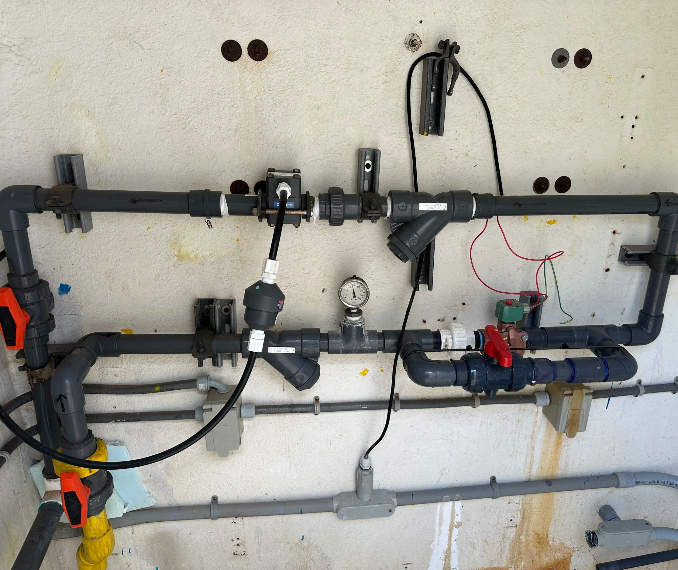 A plumbing wall with various pipes, fittings, and gauges, including black and gray PVC pipes, a pressure gauge, and electrical wiring mounted on a textured wall.