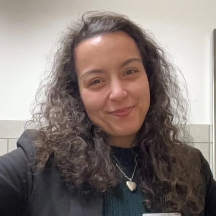 A woman with curly dark hair smiling at the camera, wearing a dark jacket and a necklace with a heart pendant.