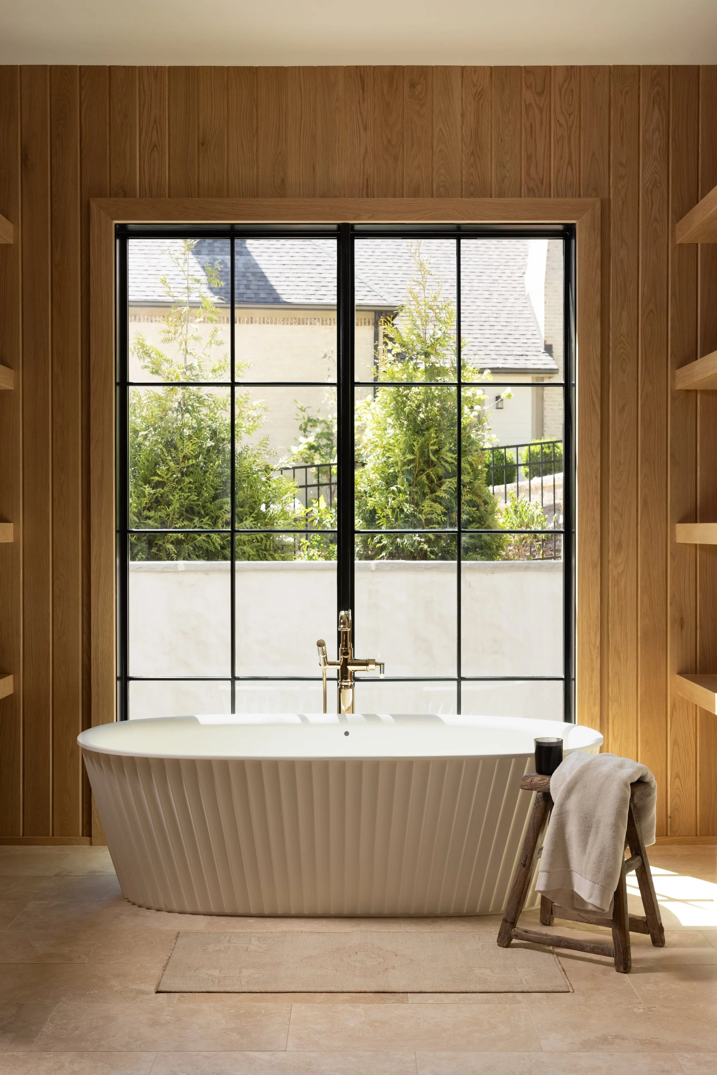 A white bathtub with ribbed sides in front of a large window with black grid panes, wooden frame and wall panels, with a view of green shrubbery and neighboring houses outside. A wooden stool holding a rolled towel and a black cup is beside the tub.
