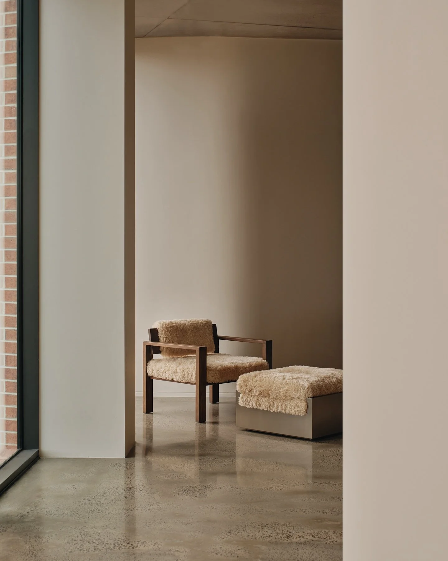 Quiet seating alcove at Programa HQ in Richmond with a timber-framed feature chair, textured cream upholstery, matching ottoman and polished concrete floor.