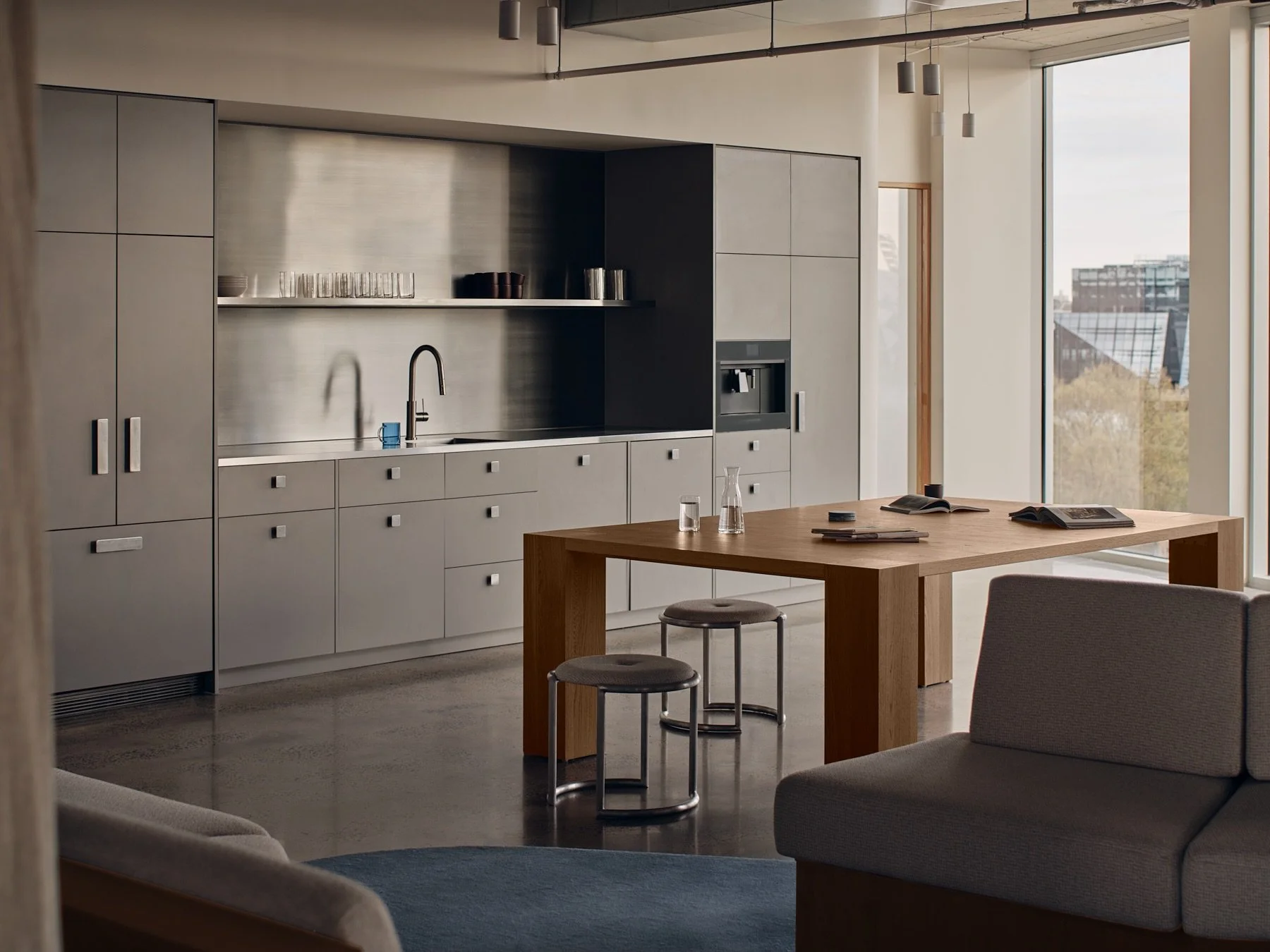 Programa HQ staff kitchen with brushed stainless steel splashback, grey cabinetry, polished concrete floors and a custom timber communal table with metal stools.