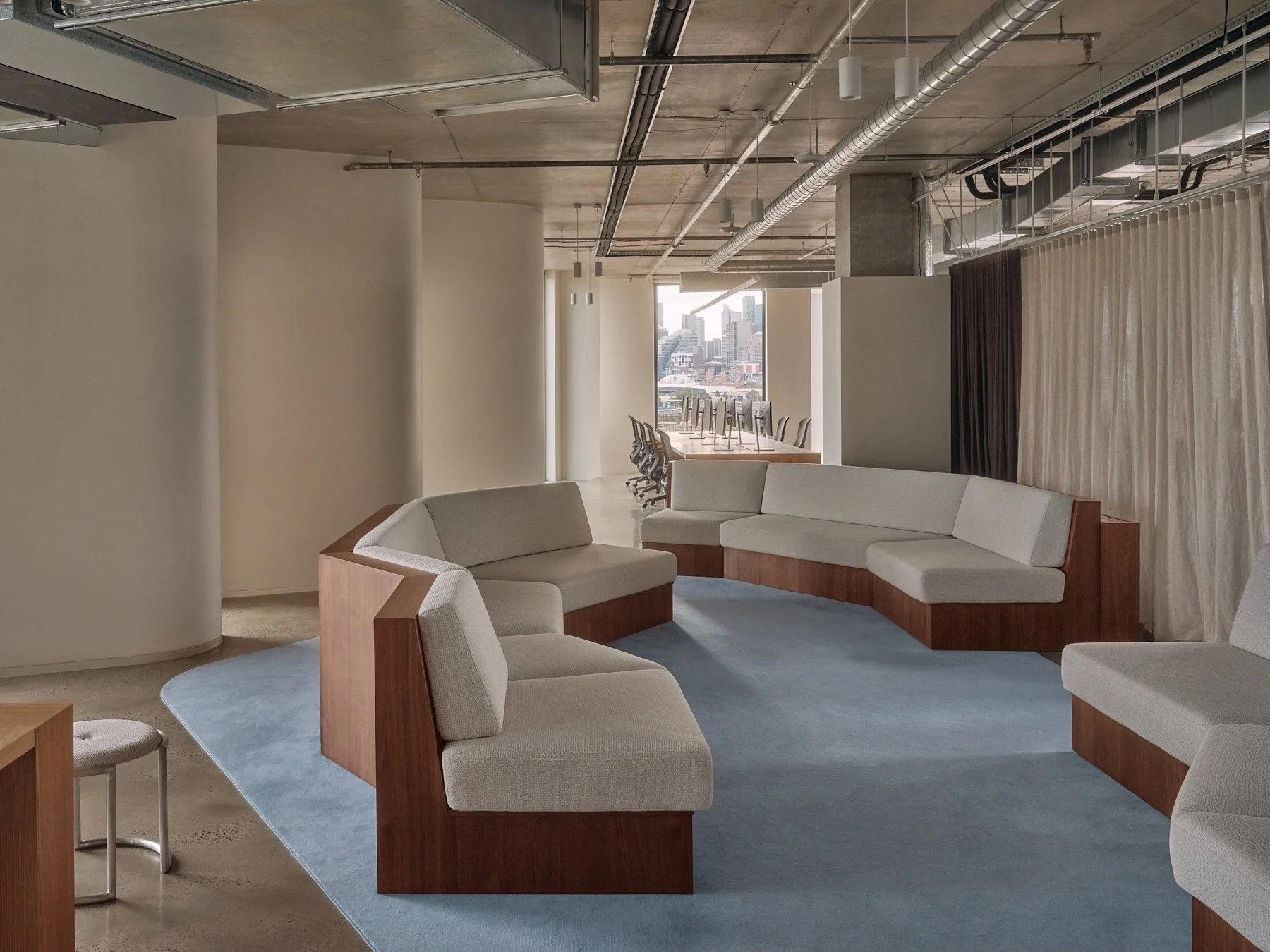 Wide view of Programa HQ's communal lounge with custom angular timber sofas, pale upholstery, blue carpet and exposed ceiling services overlooking Melbourne.