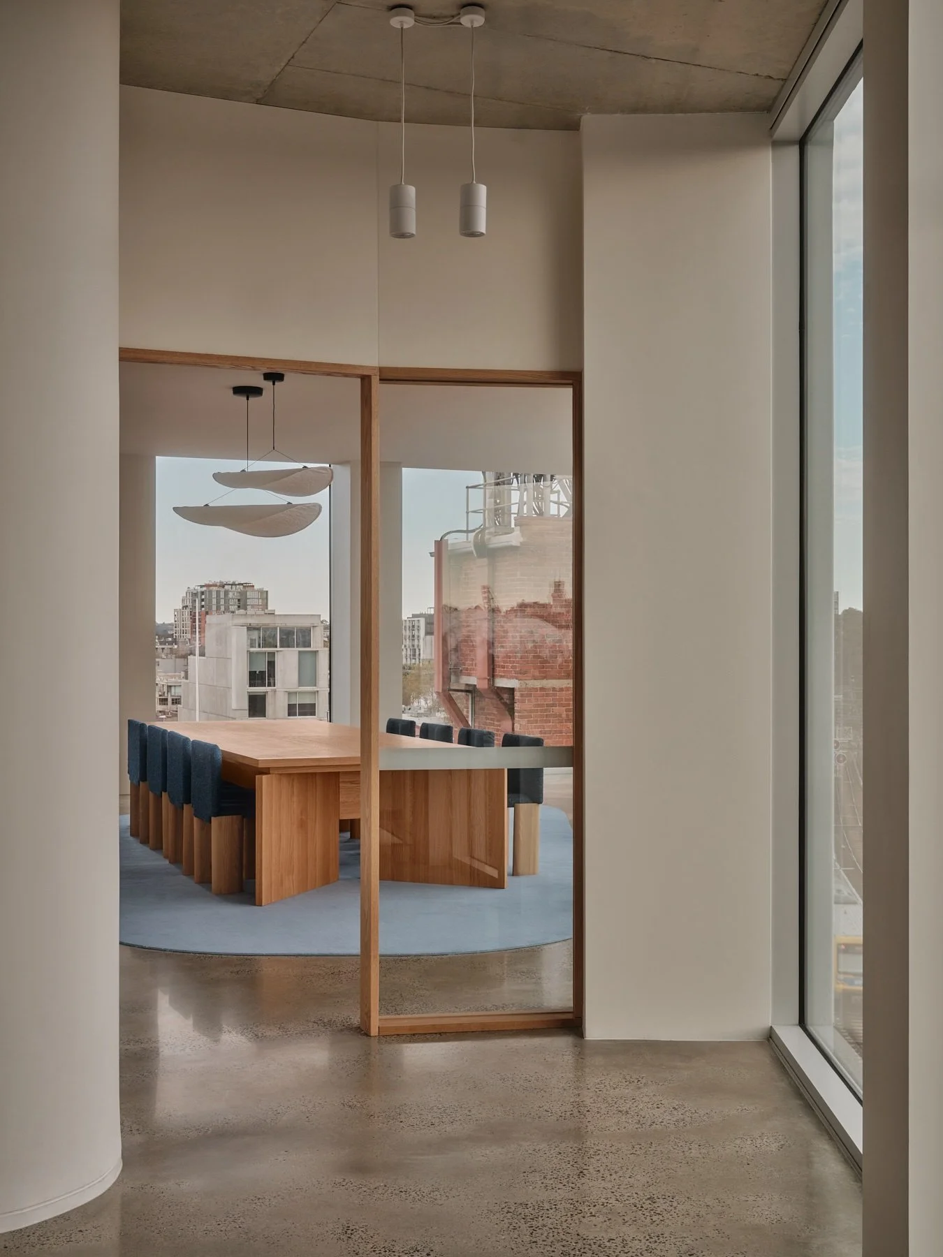 View through an American oak framed glass doorway into Programa HQ's boardroom, featuring a custom American oak conference table, blue upholstered chairs and sculptural pendants.