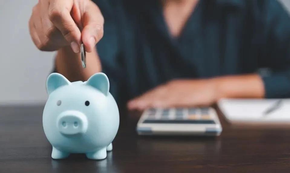 Person placing coin in blue piggy bank on table with calculator and notebook