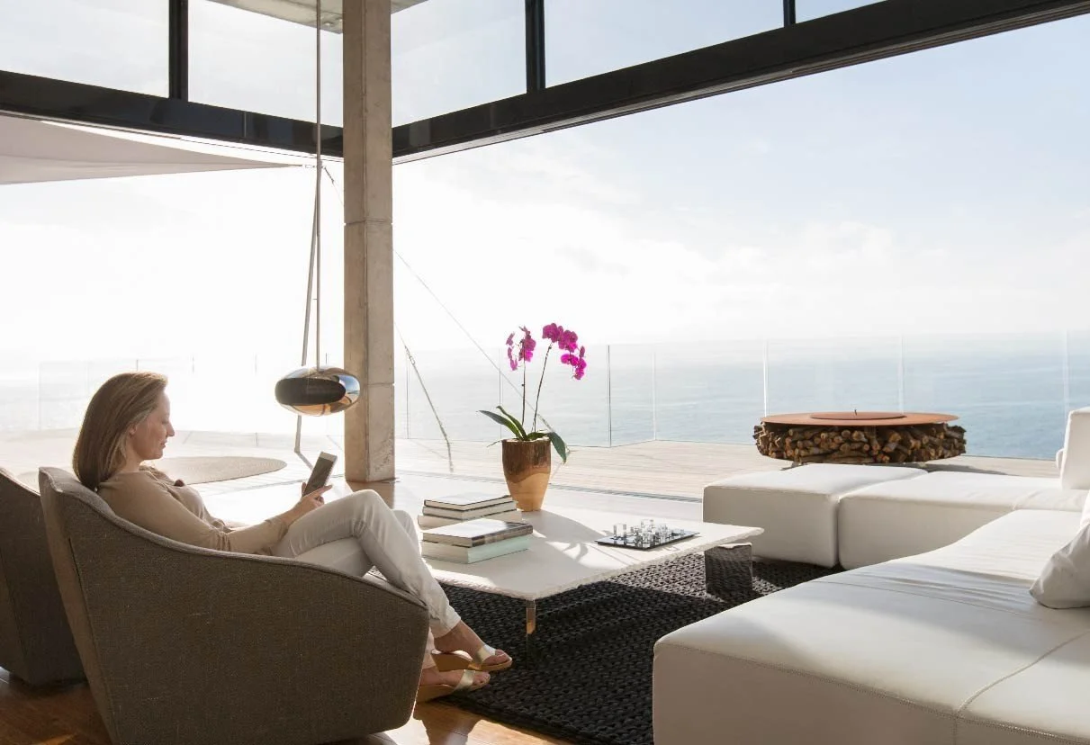 Modern living room with ocean view, featuring a woman sitting in a chair, using a tablet, white furniture, a pink orchid, and large windows.
