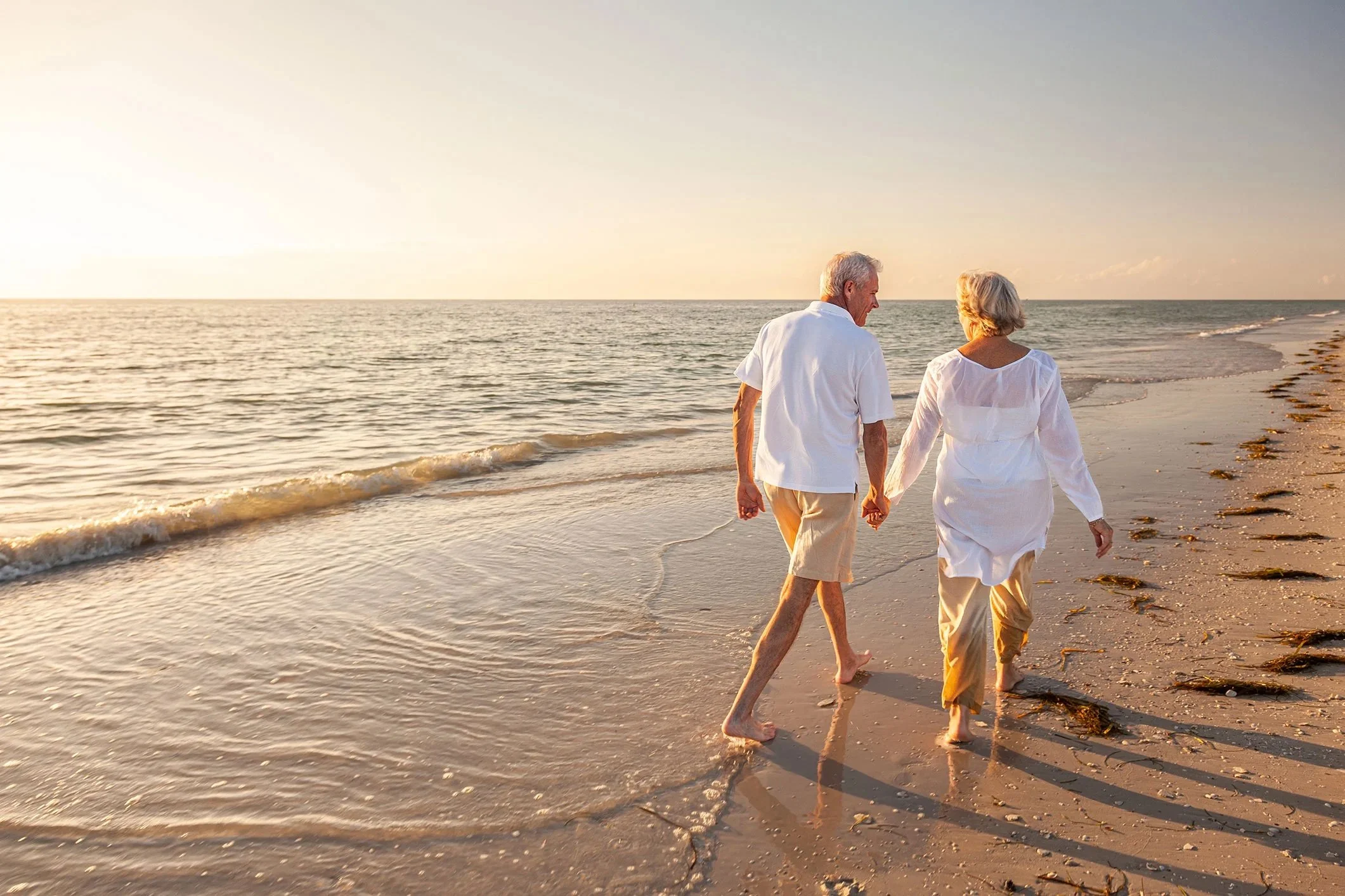 Elderly couple walking hand in hand along a sandy beach at sunset.