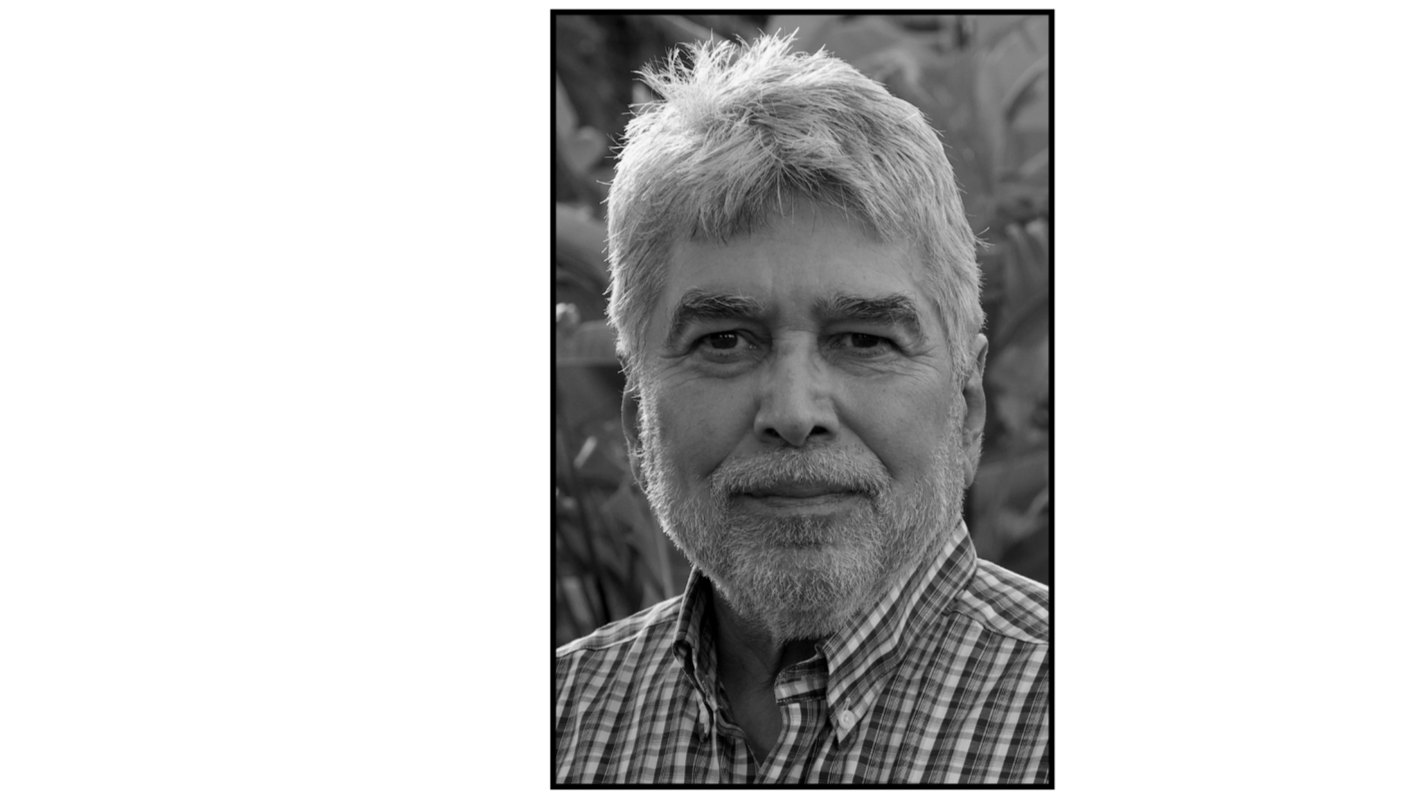 Black and white photo of an older man with gray hair and a beard, wearing a plaid shirt, looking at the camera with a slight smile.