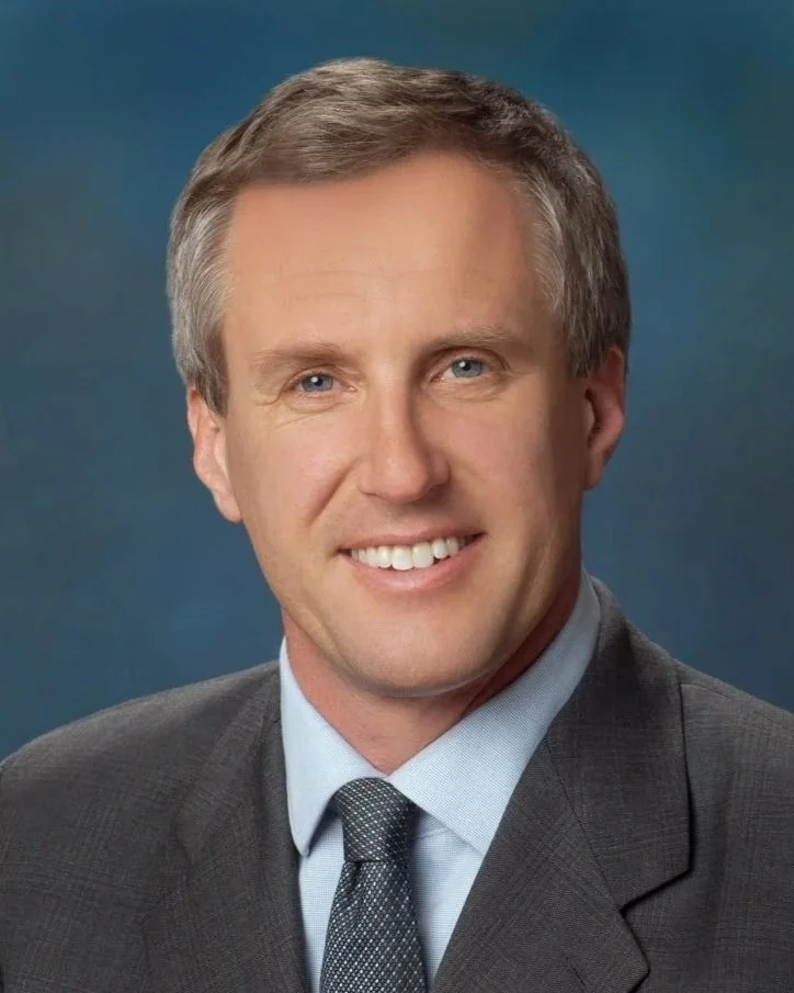 Portrait of a middle-aged man with gray hair, wearing a light blue dress shirt and a dark suit, smiling in an indoor setting.