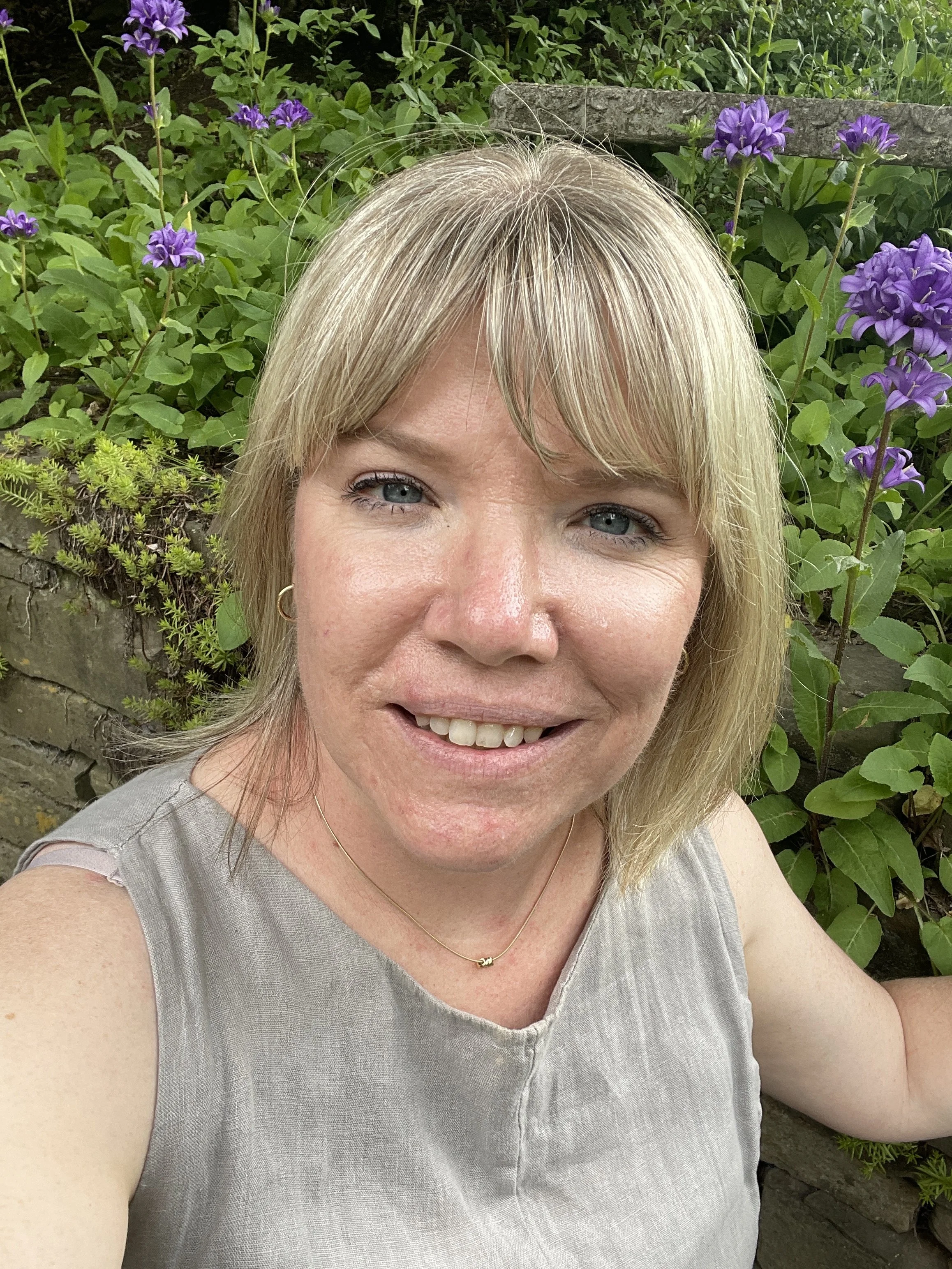 A woman with blonde hair smiling outdoors, with purple flowers and green foliage in the background.