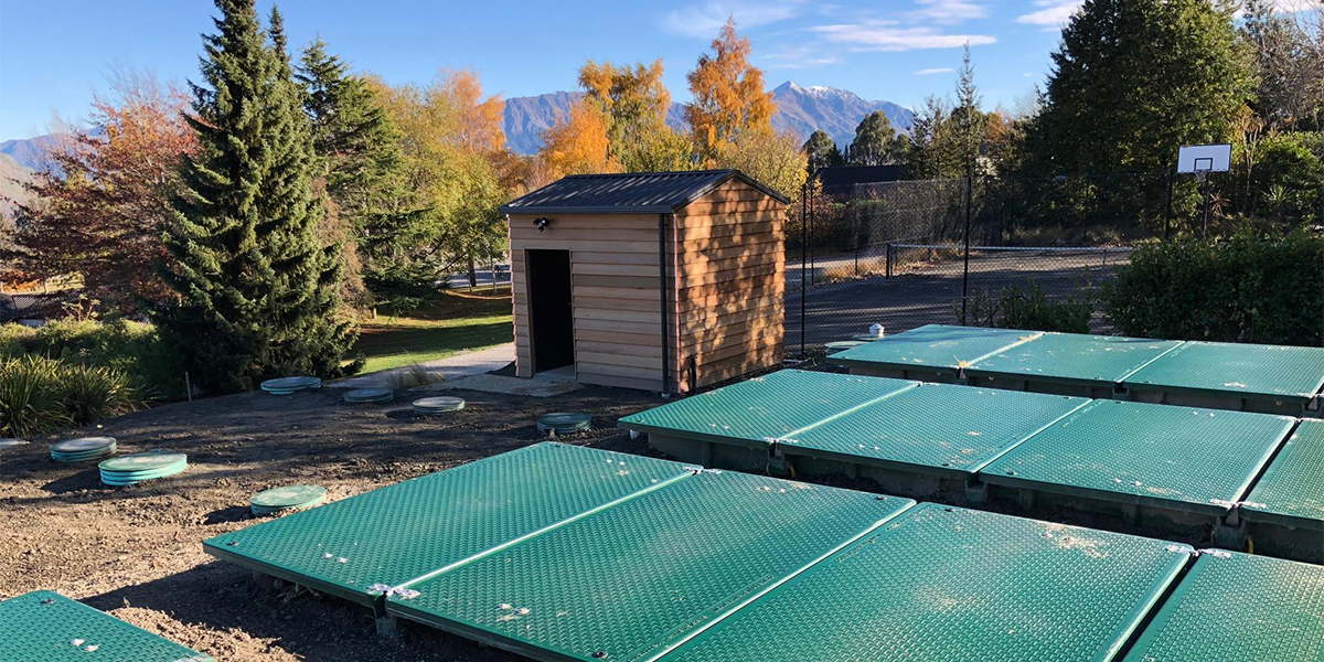 Outdoor scene with a small wooden shed, a wastewater treatment system, large evergreen trees, solar panels, a basketball court, and mountains in the background under a blue sky.
