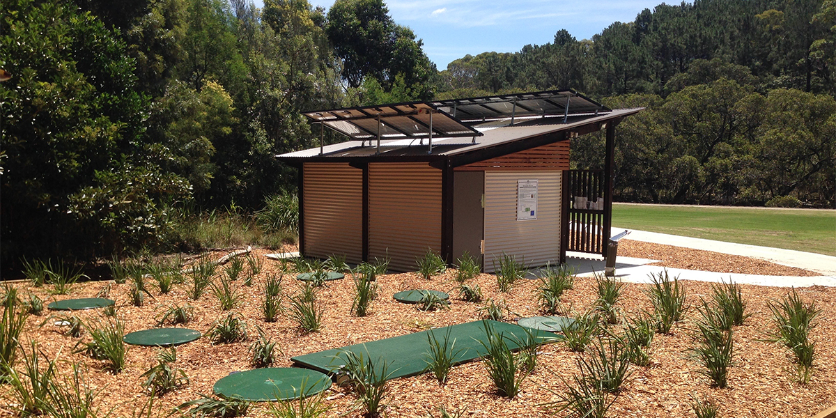 Small solar-powered structure with solar panels on roof, surrounded by young plants and mulch, in a park or garden setting.