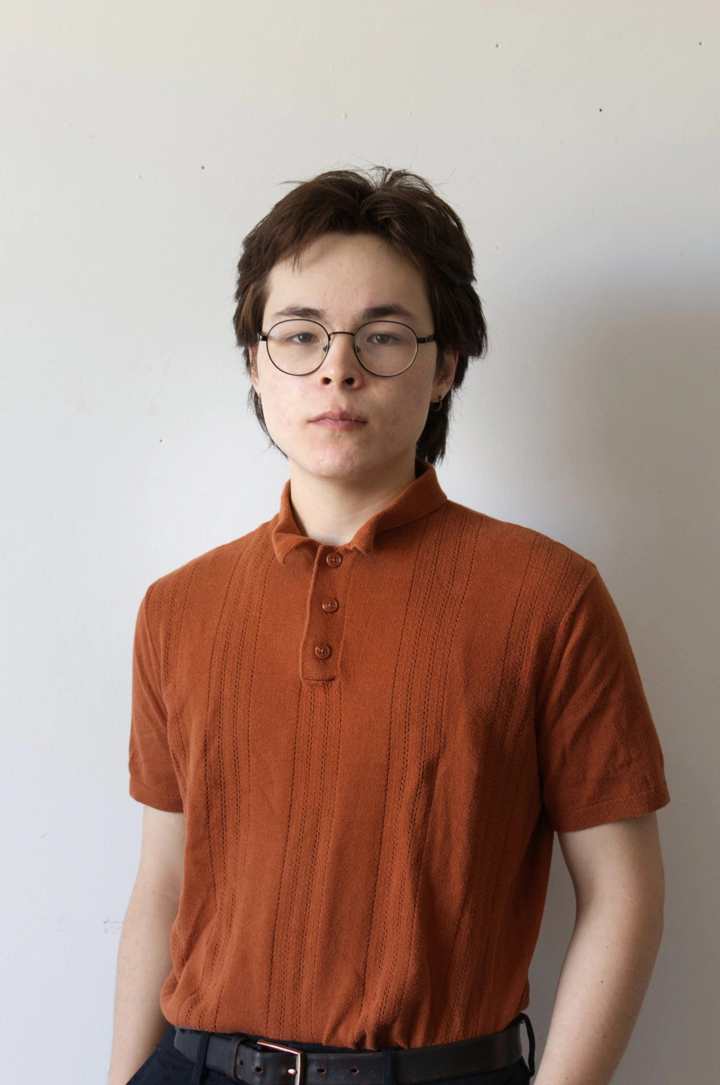 A person with short, dark, slightly messy hair wearing glasses and a burnt orange short-sleeve shirt standing against a plain white wall.