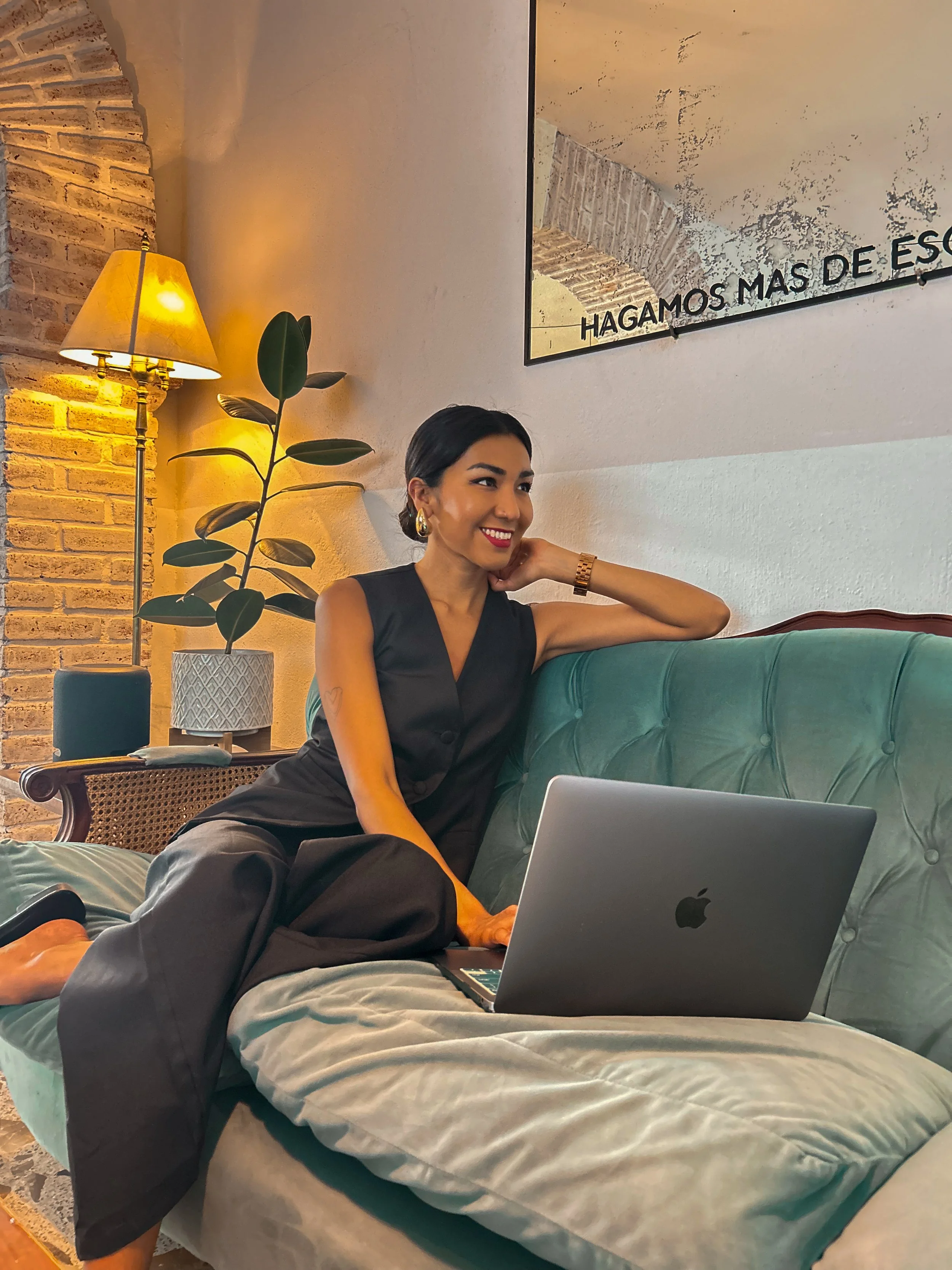 Una mujer sentada en un sofá con una laptop abreviad Apple, en una sala con lámpara de pie, planta y un cuadro en la pared, sonriendo.