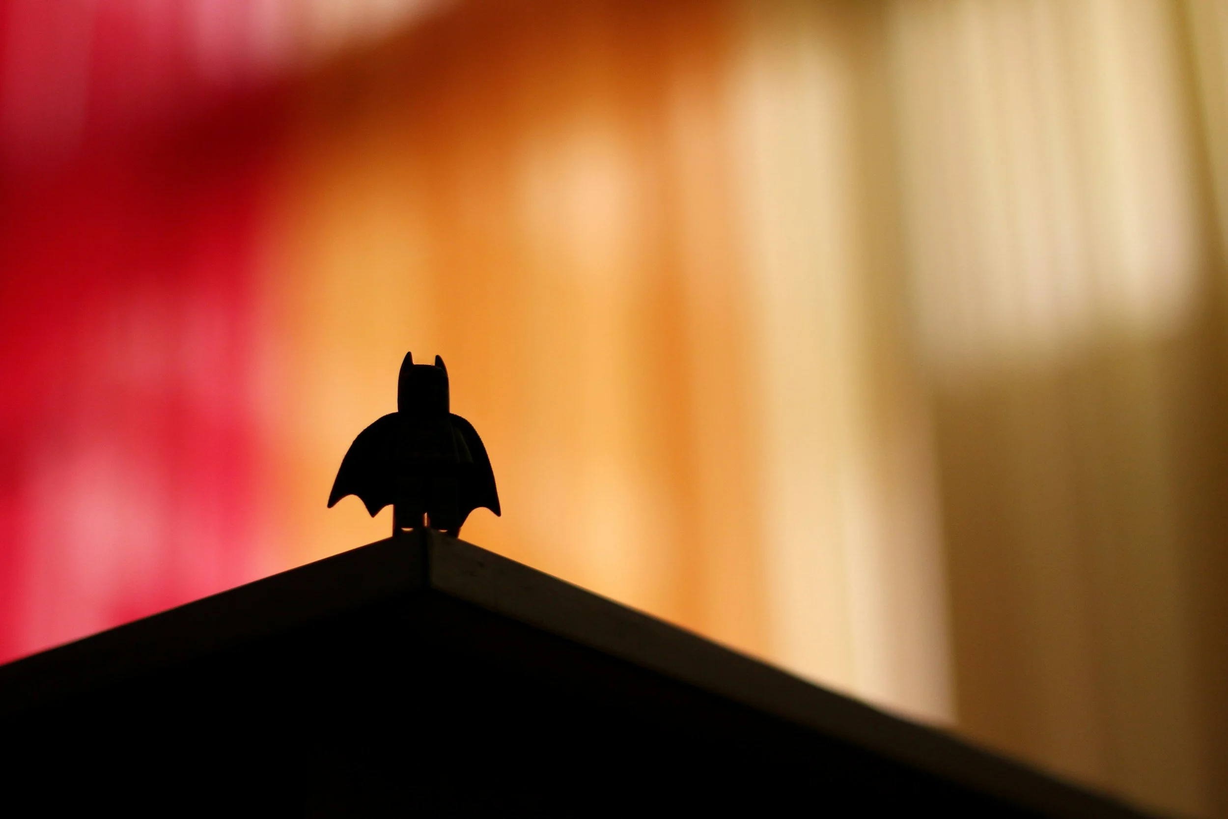 Silhouette of a small Batman figurine on the corner of a surface against a blurred colorful background.