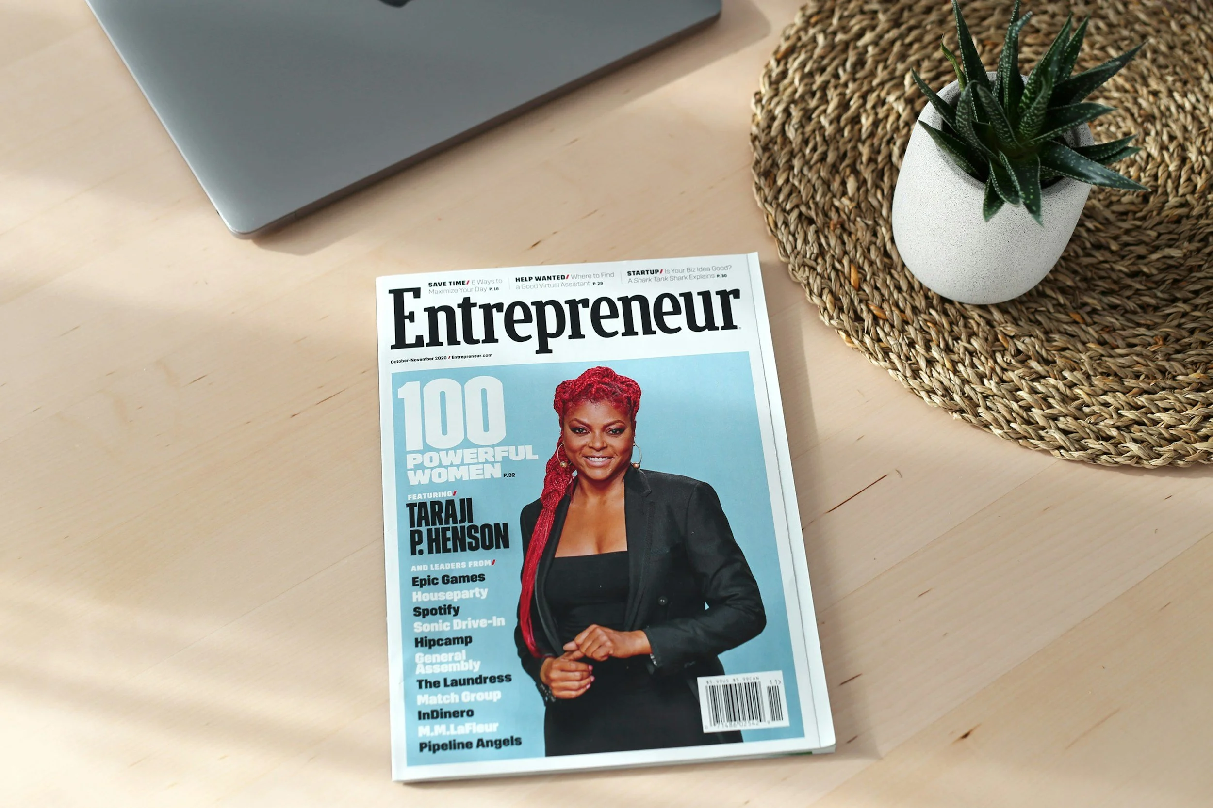 A magazine titled 'Entrepreneur' on a light wooden desk next to a laptop and a potted plant in a wicker mat. The magazine features a woman with red hair on the cover.