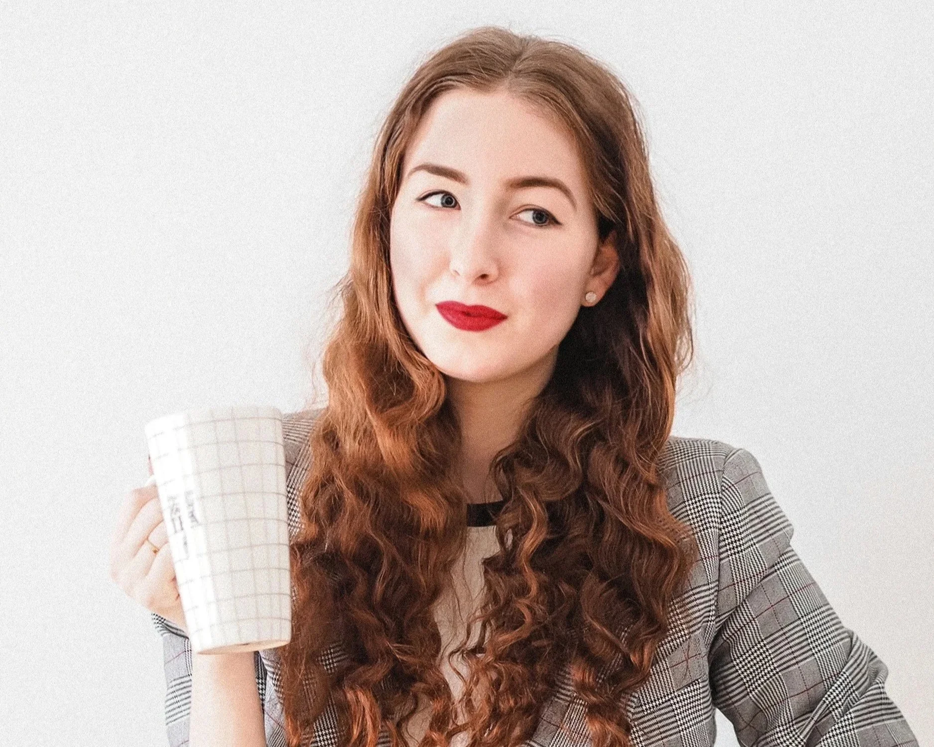 A woman with long wavy red hair holding a white mug, wearing a plaid blazer, with a neutral background.